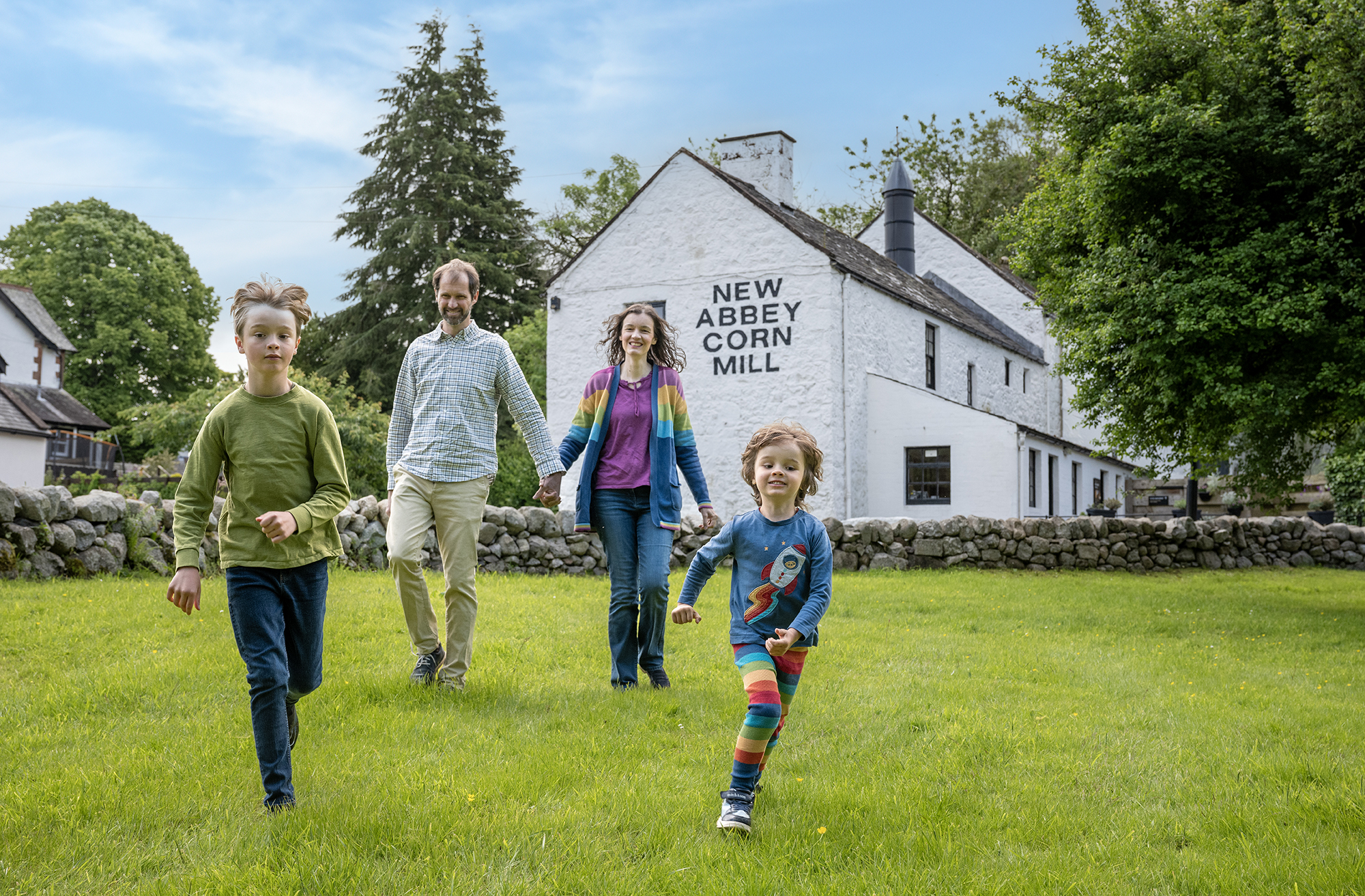 Four people, including two children, walk across a grassy field toward the camera in front of a white building with the words "New Abbey Corn Mill" painted on the wall, with trees and a stone wall in the background under a partly cloudy sky.