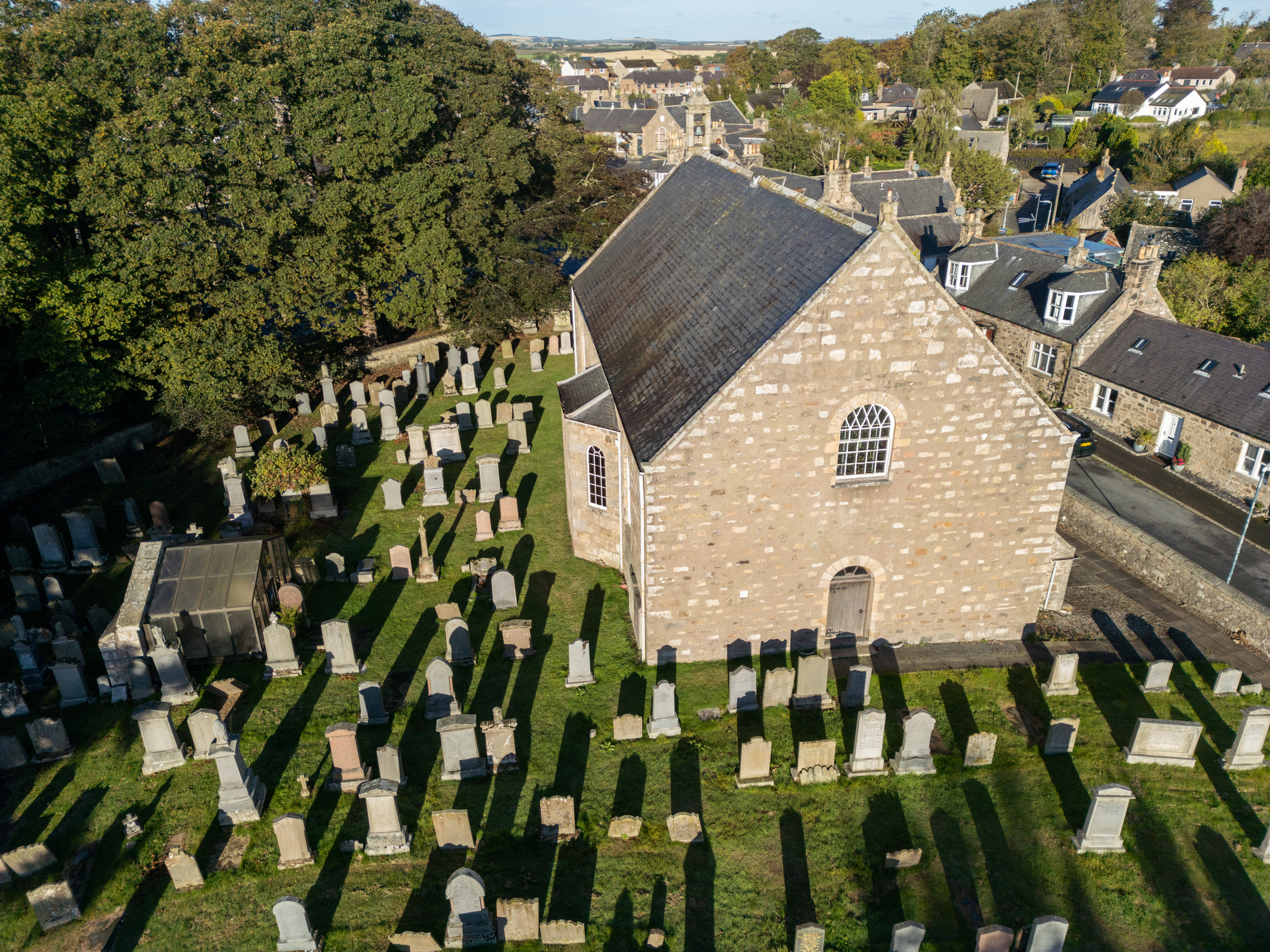 An aerial photo of a modest stone church building surrounded by gravestones. A small shelter in the churchyard protects a medieval tomb from the elements. 