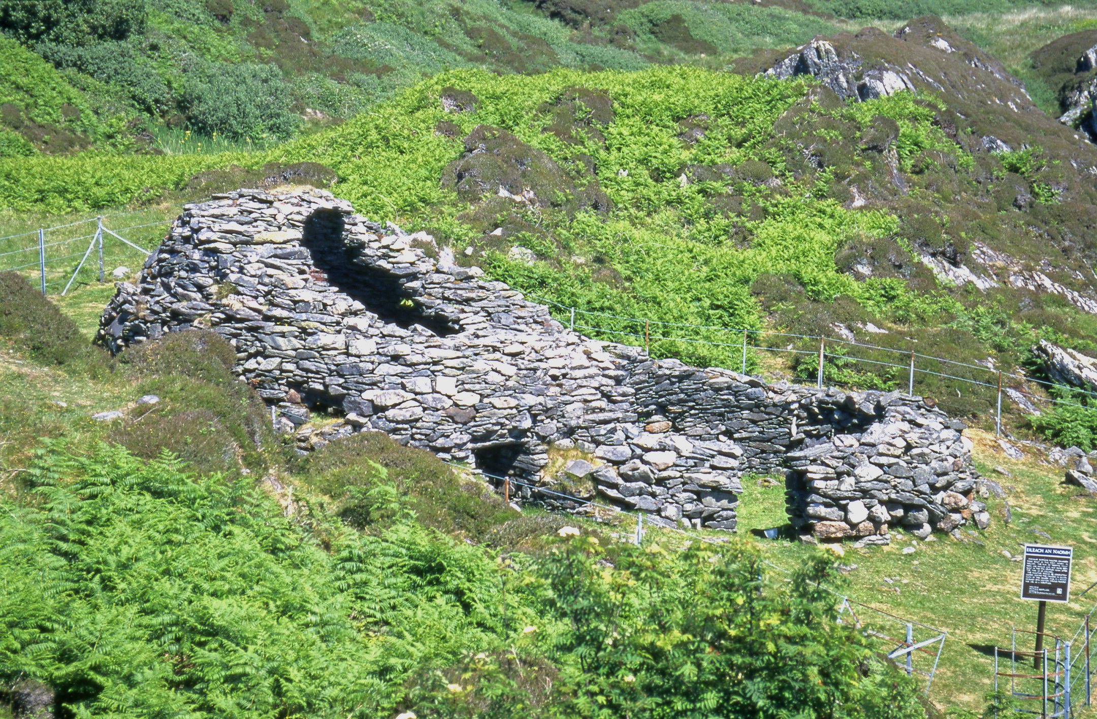 A stone beehive-shaped structure with a curved roof and small entrance, surrounded by green vegetation and rocky hills.