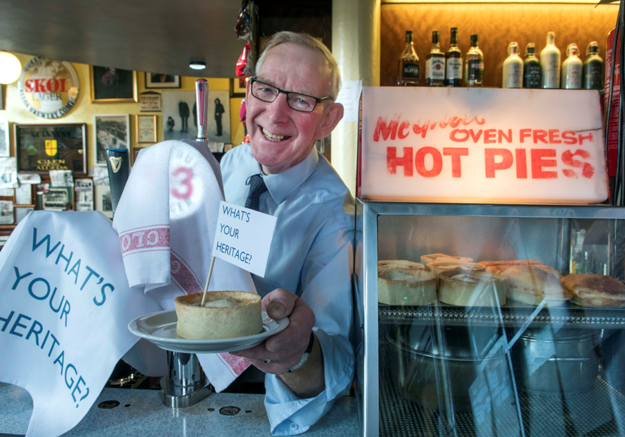 Older man, wearing glasses and a blue button up shirt and tie, stands behind a bar holding out a pie with a flag in it that reads 'What's your heritage'.