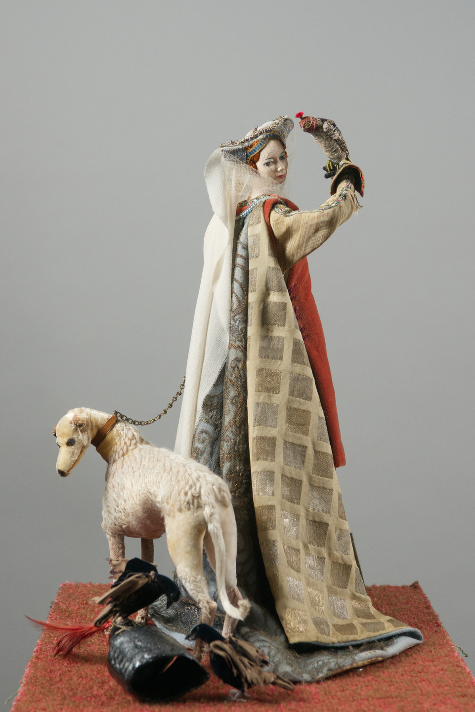 A detailed model of a medieval woman wearing layered embroidered robes and a long veil, standing with one arm raised to hold a falcon. A light‑coloured hound on a leash stands at their side, with feathers and hunting gear arranged on the ground beneath them.