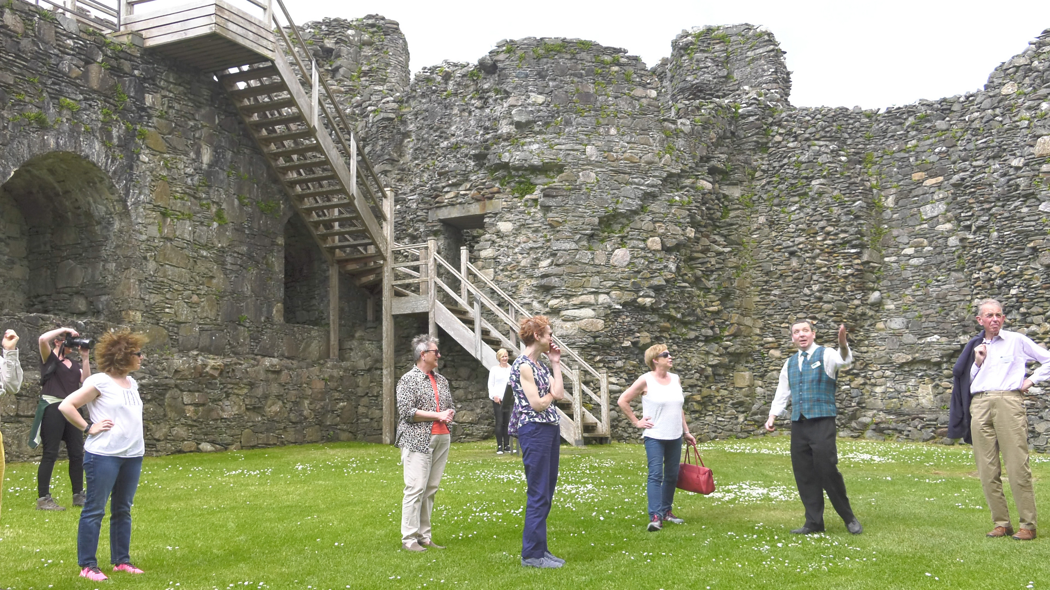 A group of visitors standing on a grassy area inside a stone castle courtyard, listening to a guide near wooden stairs leading to an upper level.
