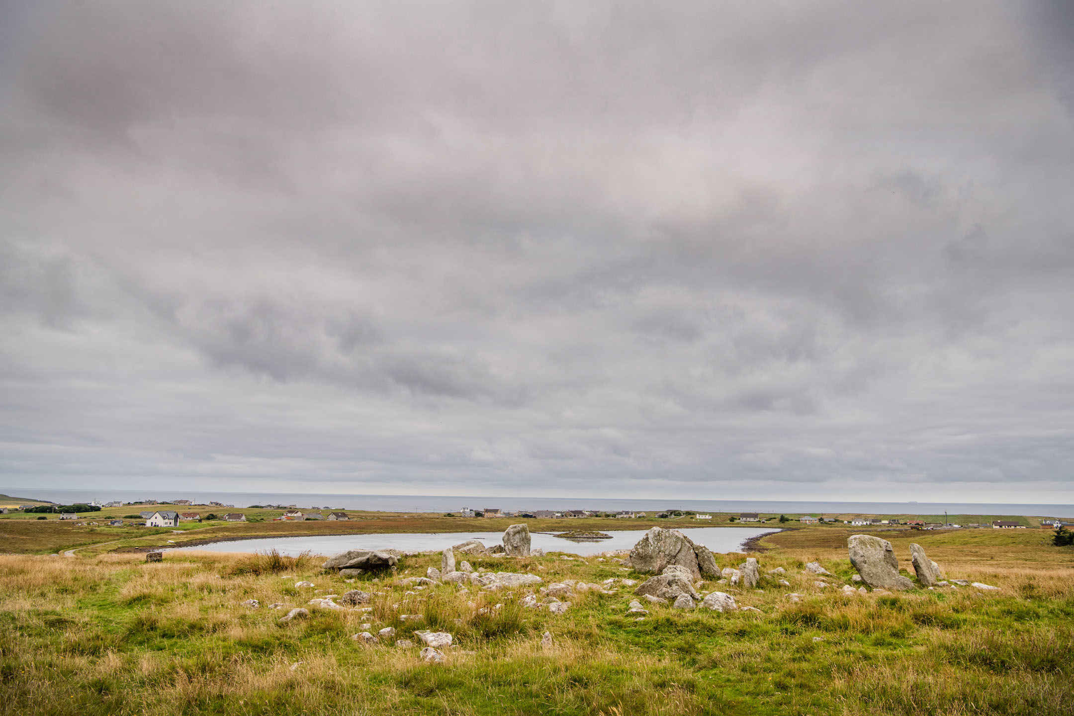 Panoramic view of stone circle remains with loch and scattered houses under grey clouds.