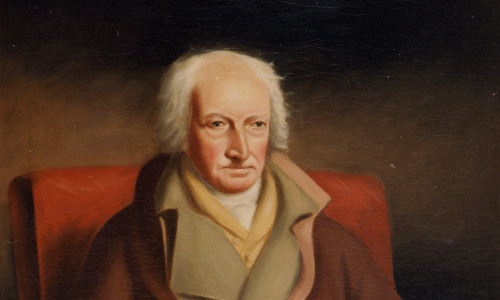 Image is a colour portrait painted of a person sitting in a chair with their hands in their lap, fingers interlaced. They are wearing several layers with a brown coat, and are looking with a serious expression to the right of the painter.