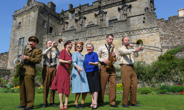  A group of six performers in 1940s style clothes stand in the gardens in front of Stirling Castle. Four band members play brass instruments as they stand either side of three women in colourful dresses.