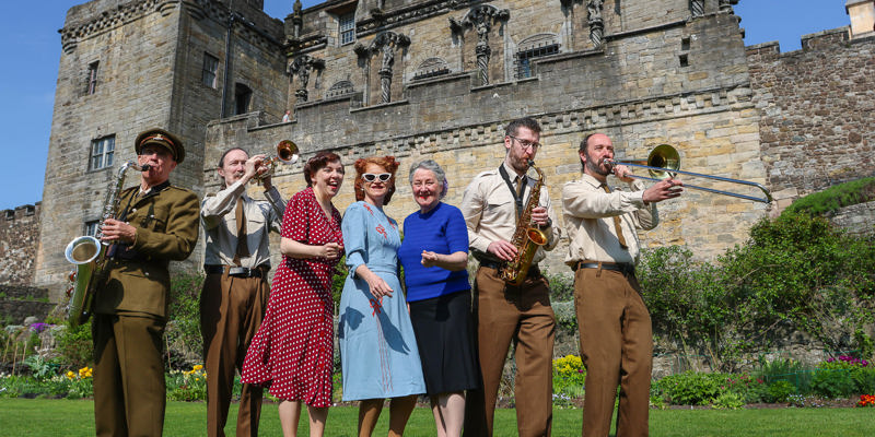  A group of six performers in 1940s style clothes stand in the gardens in front of Stirling Castle. Four band members play brass instruments as they stand either side of three women in colourful dresses.