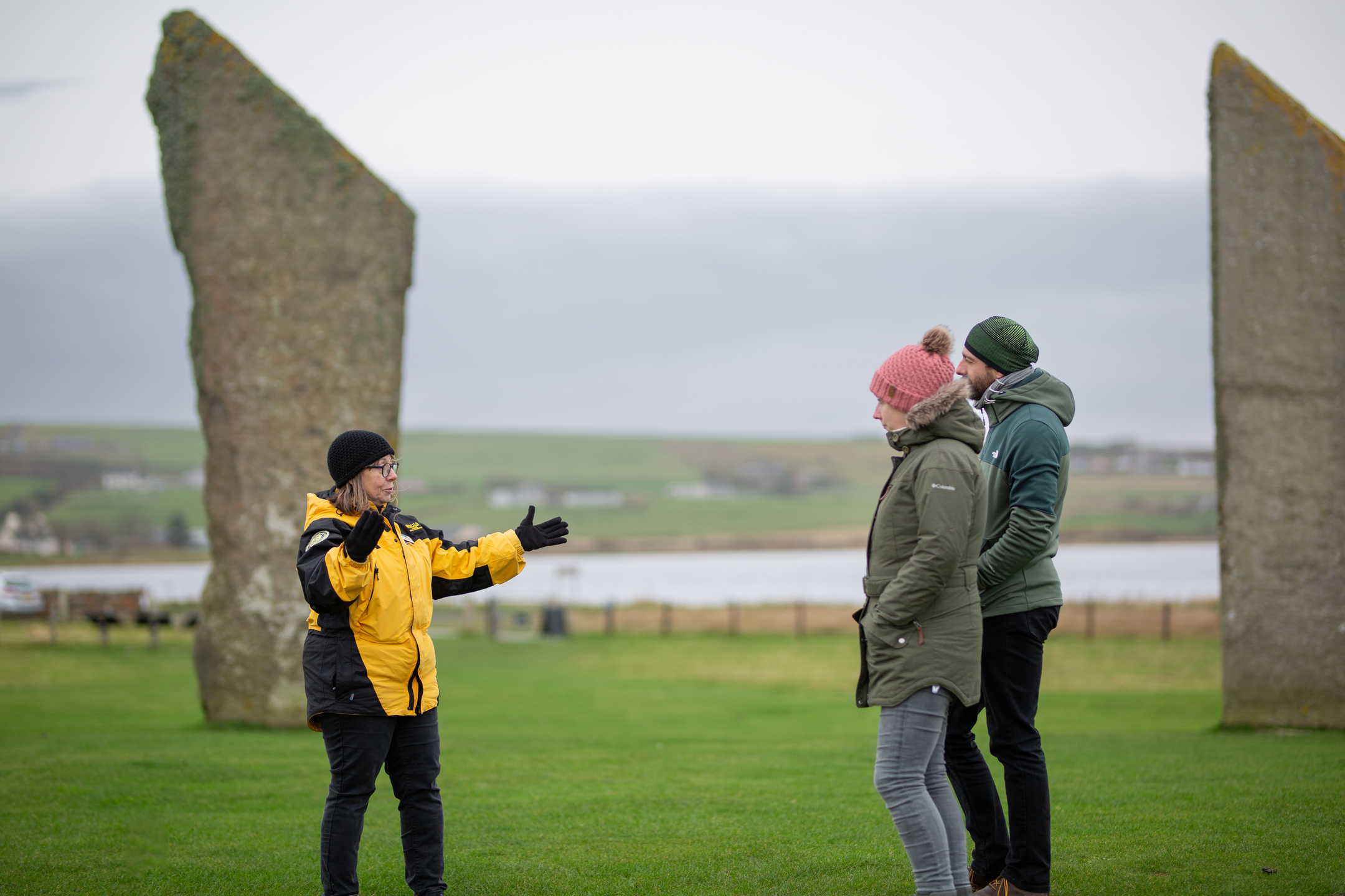 An Orkney ranger in a yellow jacket taking to two visitors stood between two large neolithic standing stones