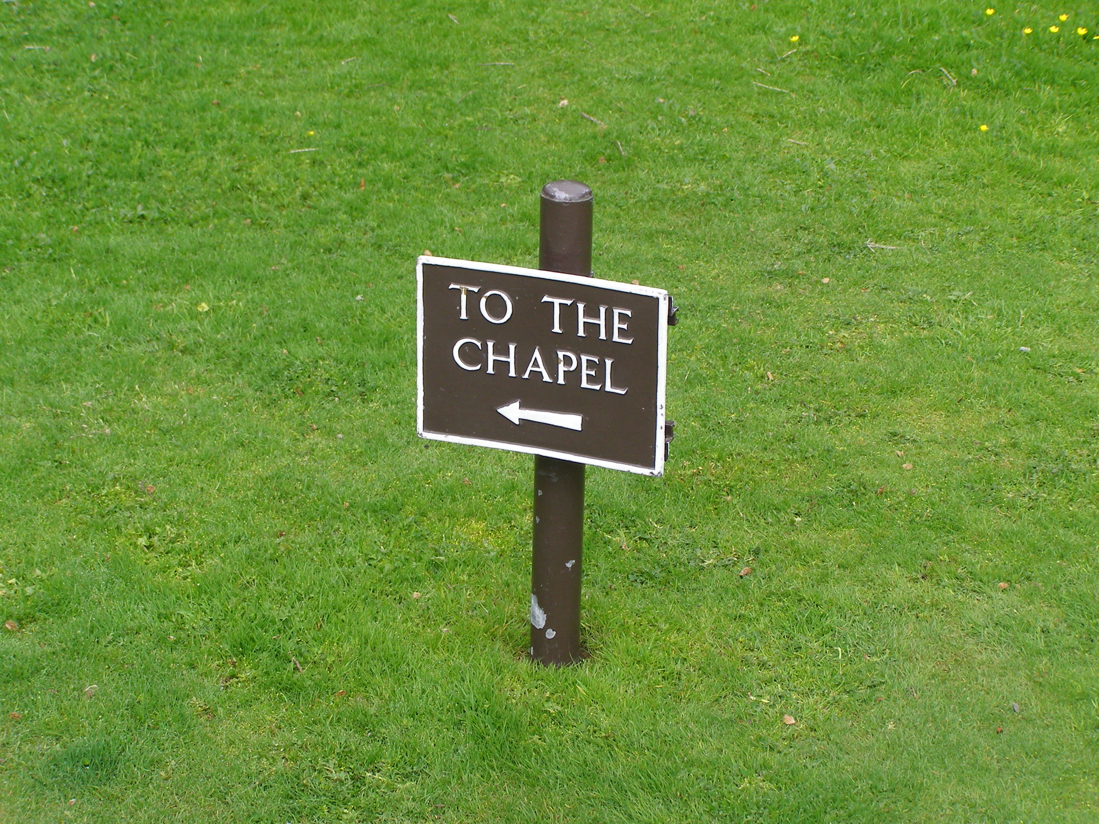A small brown sign on a grassy lawn reading “To the Chapel,” with an arrow pointing left.