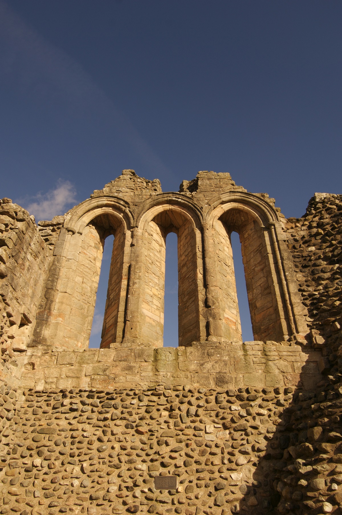 Close-up view of a tall stone wall from a ruined castle, featuring three narrow arched window openings side by side. The weathered stonework is set against a clear blue sky.