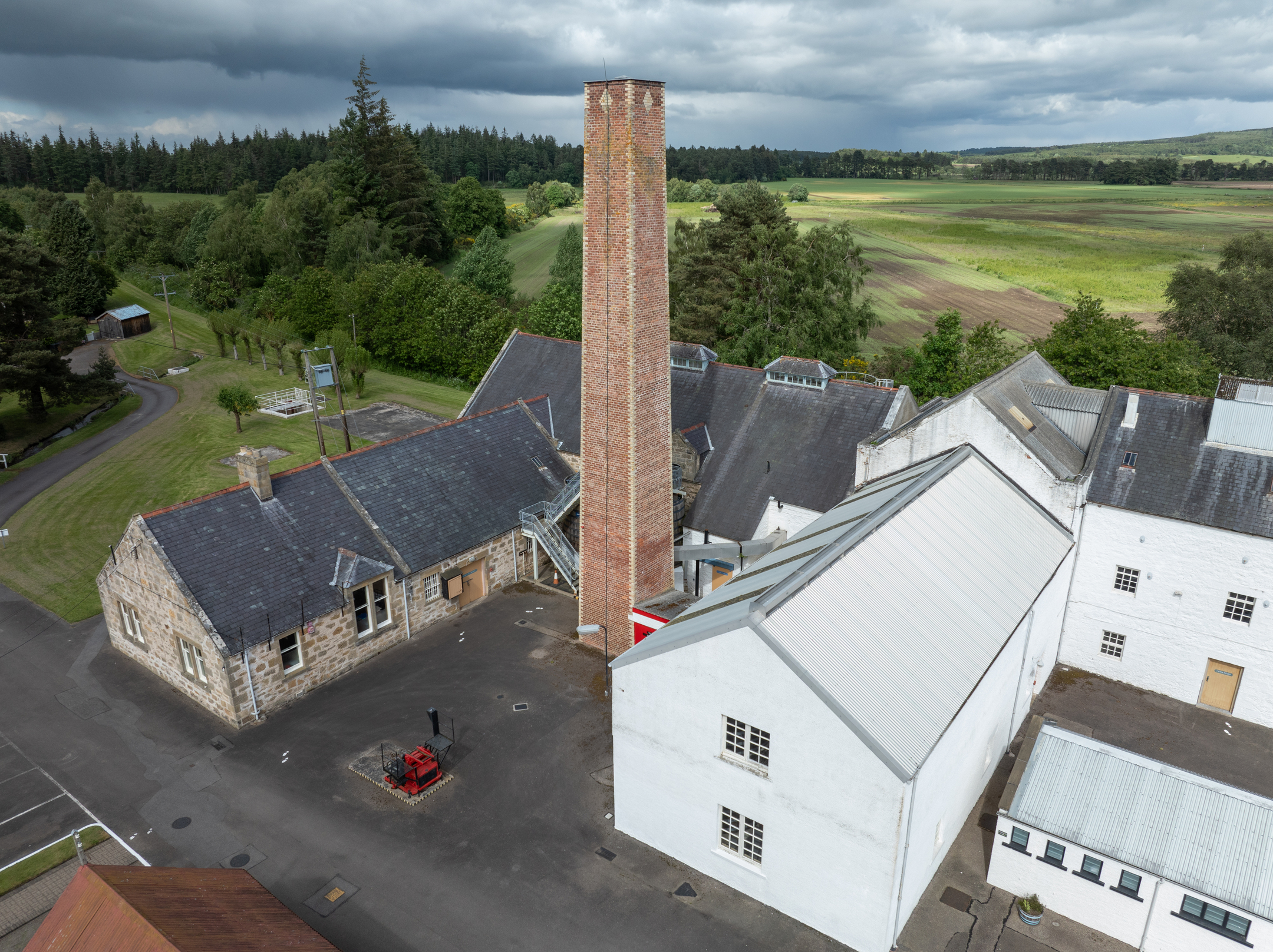 Aerial view of Dallas Dhu distillery centred on a tall red-brick chimney rising above whitewashed and stone buildings.