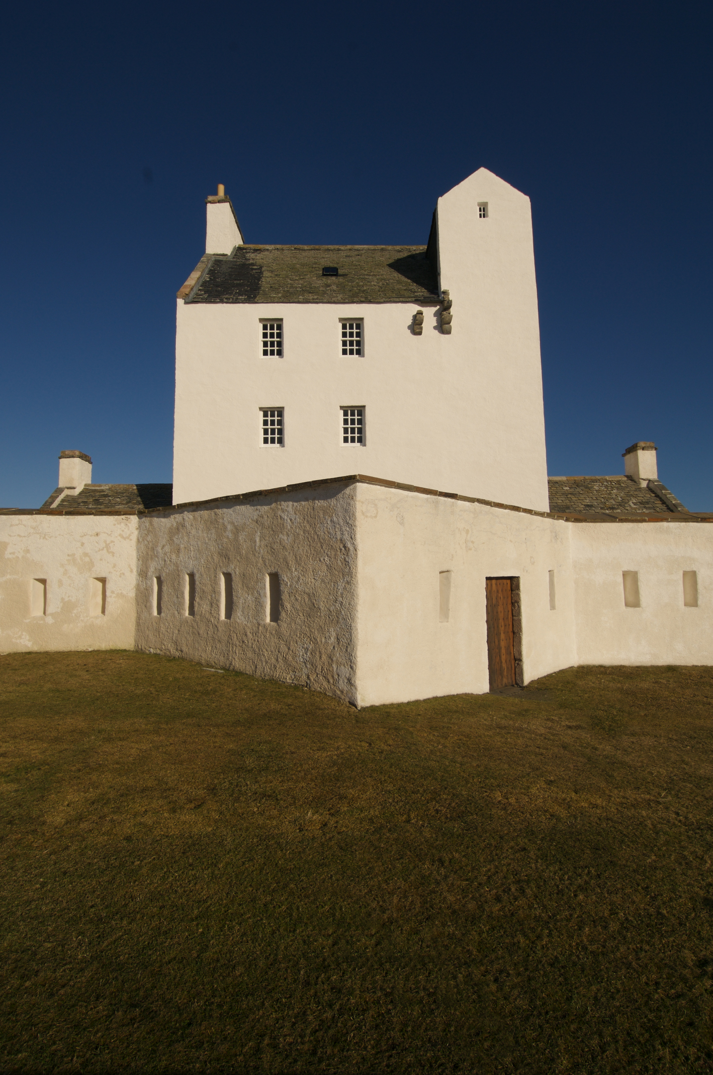 Front-facing view of Corgarff Castle’s white tower and defensive wall under a deep blue sky, with grassy ground in front.