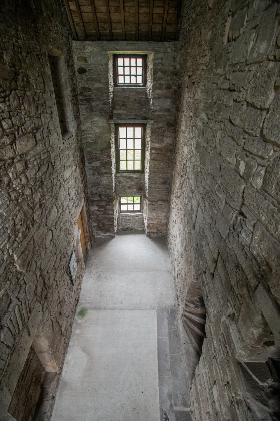 A high view of the interior of a narrow, stone room that looks historic. There are three windows of varying heights, with light coming through them. The floor is light grey. 
