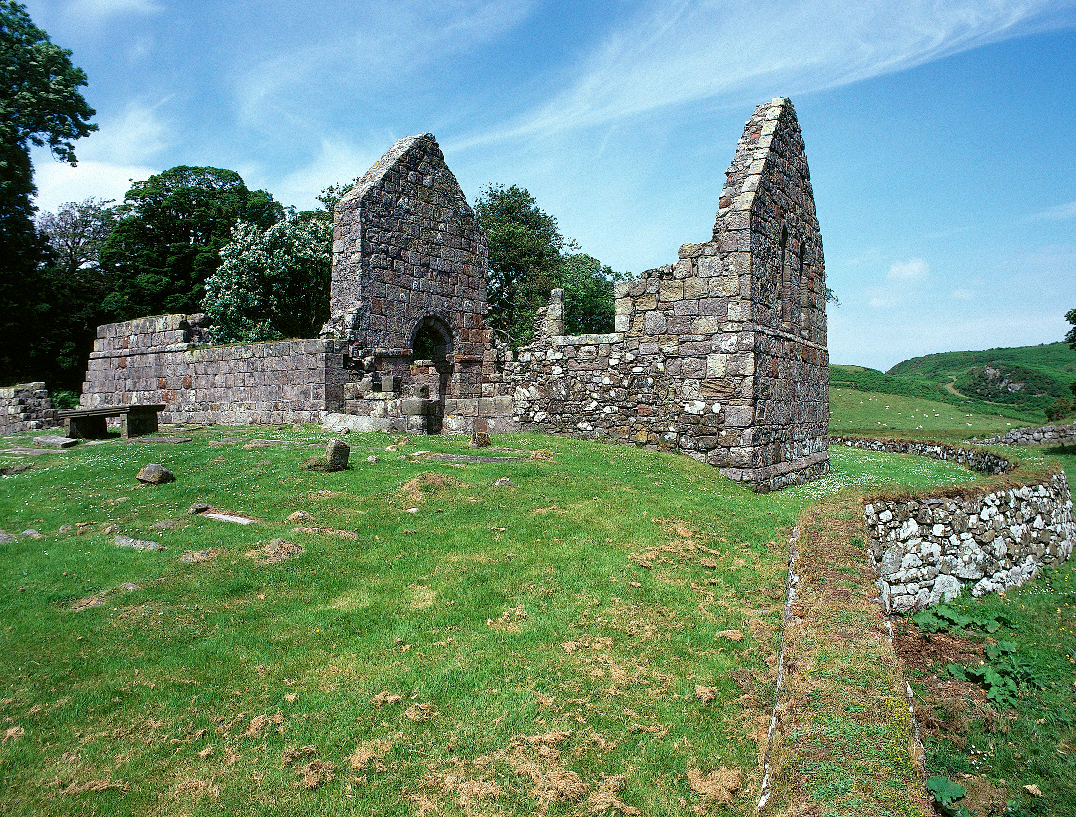 Detailed view of St Blane’s Church ruins showing two tall gable ends with arched openings and sections of stone walls. The structure stands on a grassy area with scattered stone fragments and a low boundary wall. Behind the ruins, trees provide shade, and rolling hills stretch into the distance under a bright sky with thin clouds.