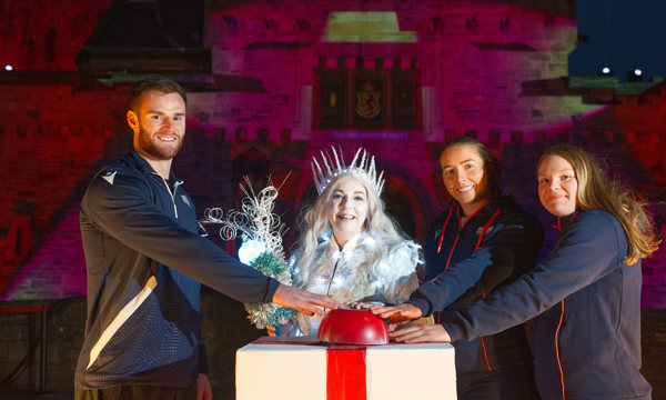A woman dressed in a silver dress with lights wearing a white crown stands in front of a white and red podium. On her left is a man dressed in a black sports jumper and trousers, and on her right are two women dressed in Edinburgh Rugby jumpers. They are all pressing a big red button. Behind them is a stone castle lit up with red, purple and green colours