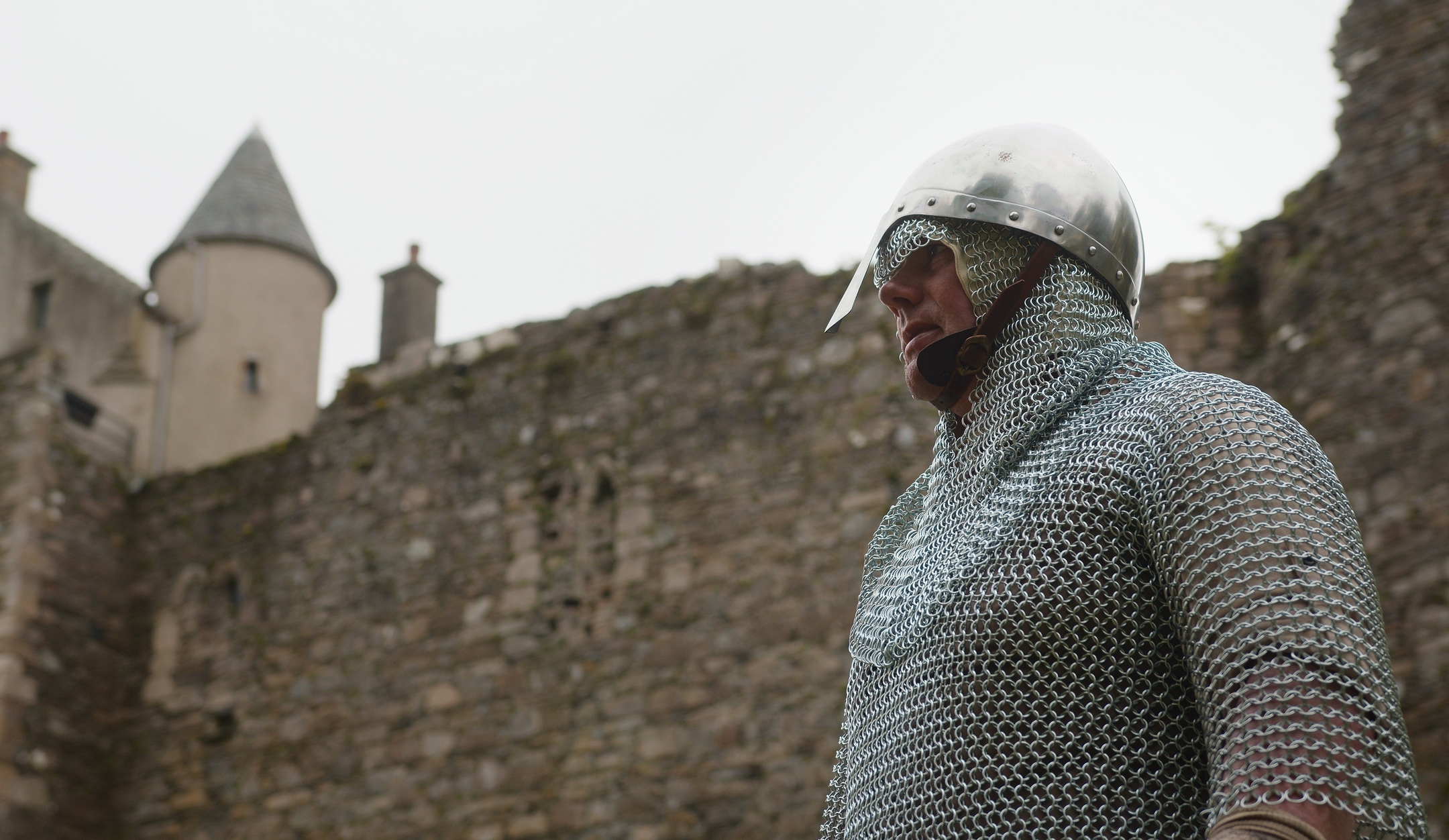 A person in chainmail armour and a metal helmet standing in a medieval stone courtyard, with castle walls rising behind them.