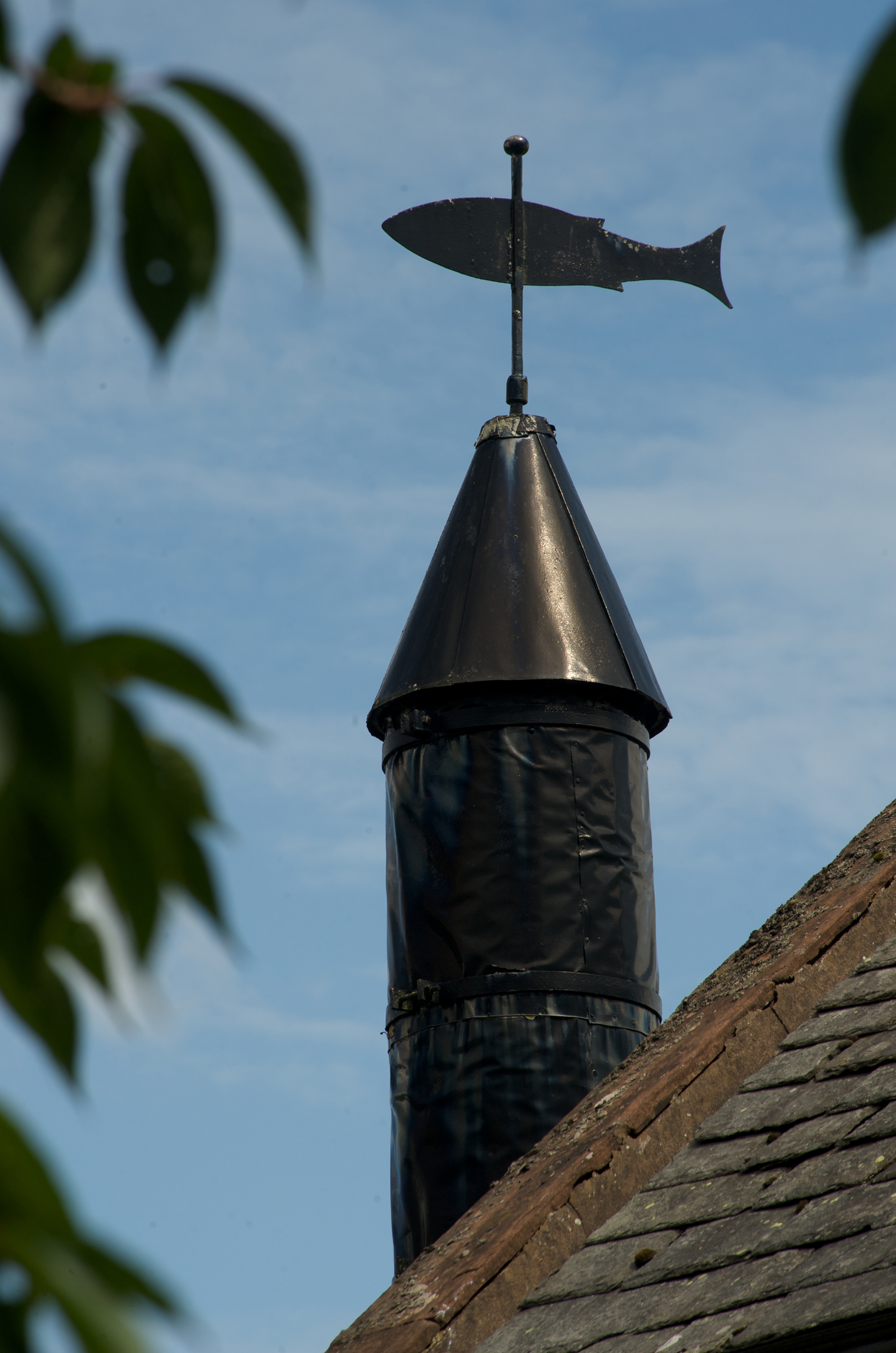 A close-up of a metal weather vane shaped like a fish, mounted on top of a conical roof vent against a blue sky.
