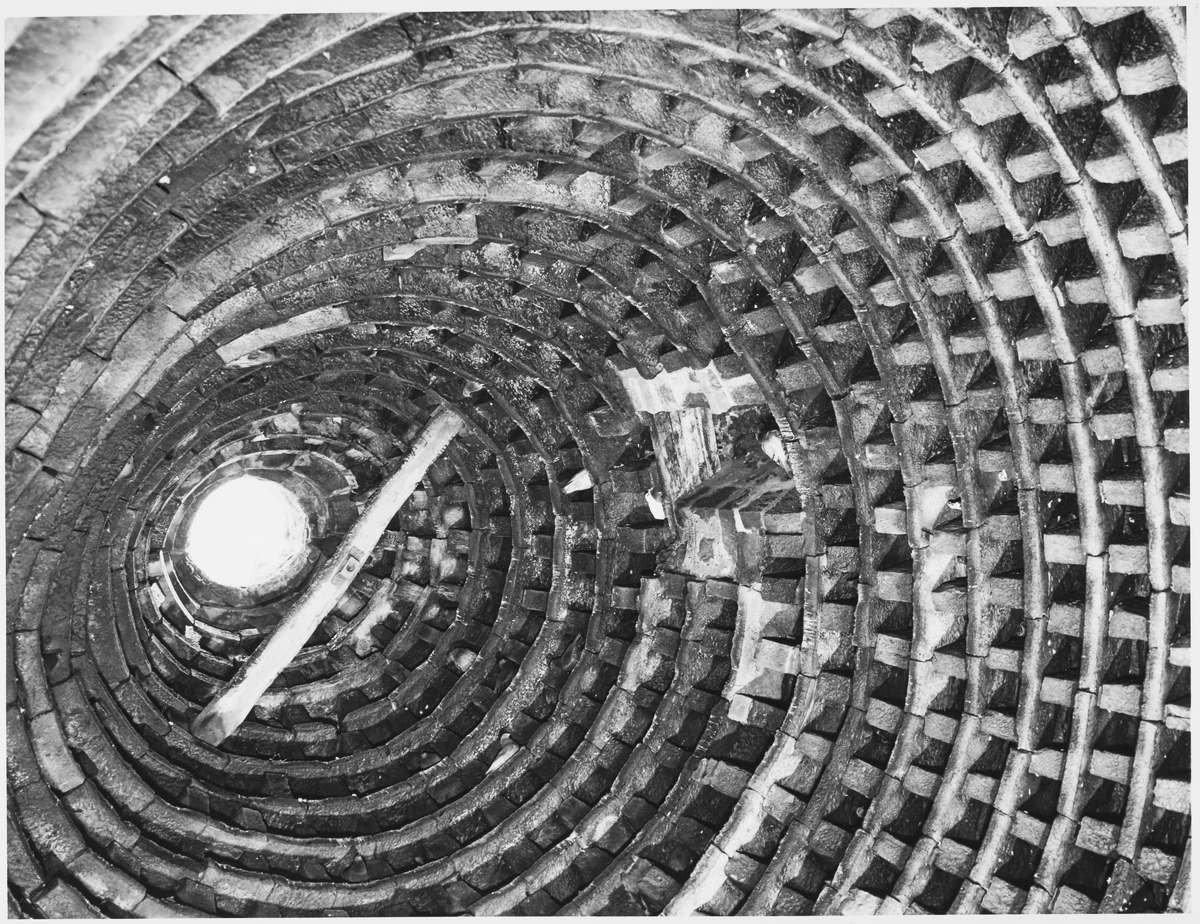 A black‑and‑white view looking upward inside a circular brick structure made of many concentric rings. The bricks are arranged in a repeating, lattice‑like pattern, creating a spiralling effect toward an opening of bright light at the top. A long wooden beam crosses diagonally through the centre of the frame