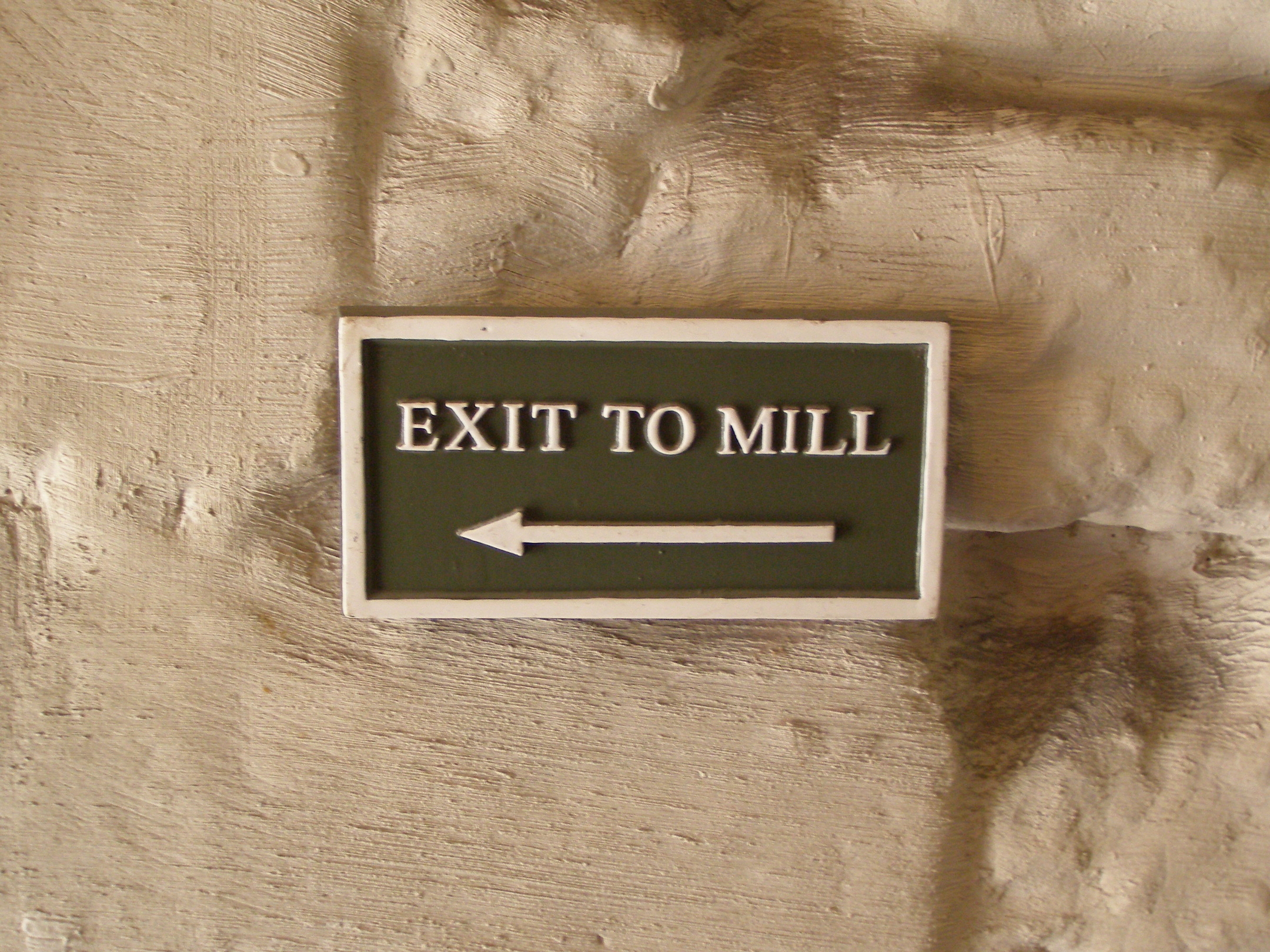 A sign reading “Exit to mill” with an arrow pointing left, mounted on a textured stone wall.