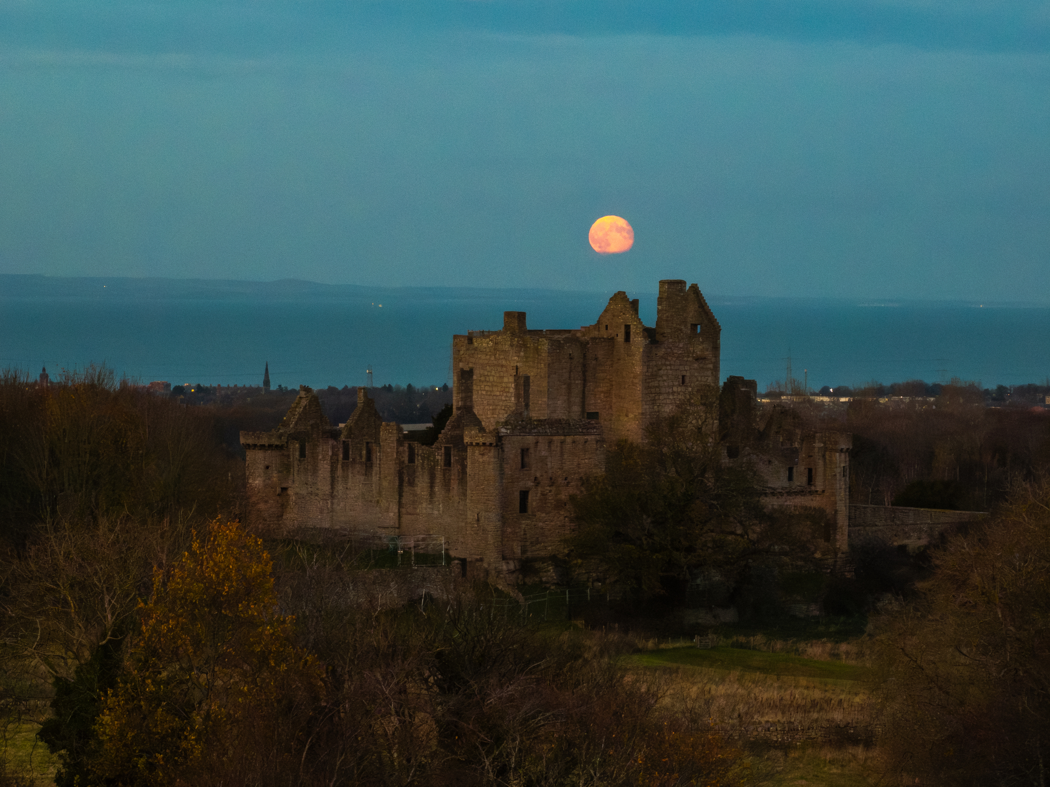 Craigmillar Castle illuminated by low evening light as a full moon appears above the horizon.