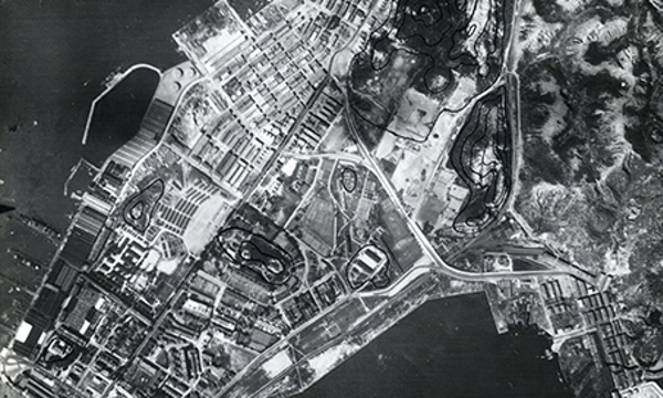 A black and white aerial photograph of Hung Hom Wan, Hong Kong. To the bottom of the image is the sea, with a winding coast above. Buildings and vast fields are visible on the land