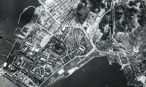 A black and white aerial photograph of Hung Hom Wan, Hong Kong. To the bottom of the image is the sea, with a winding coast above. Buildings and vast fields are visible on the land