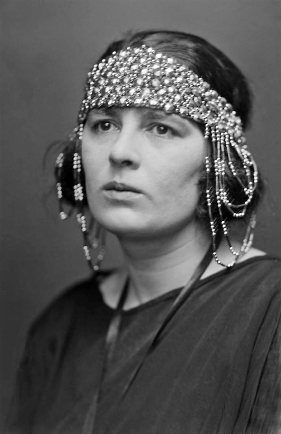 Black and white photograph of a person from the shoulders up. They are wearing a dark coloured top and an elaborate beaded headdress that runs across the forehead and falls down over both ears.