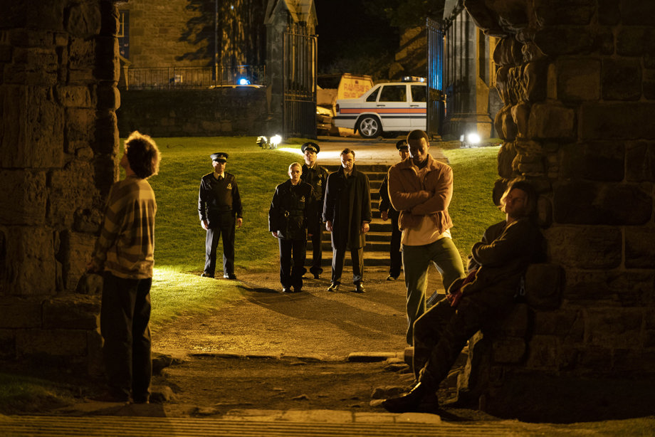 Four police officers and a DS in cathedral grounds in the night walking towards three young men looking uneasy