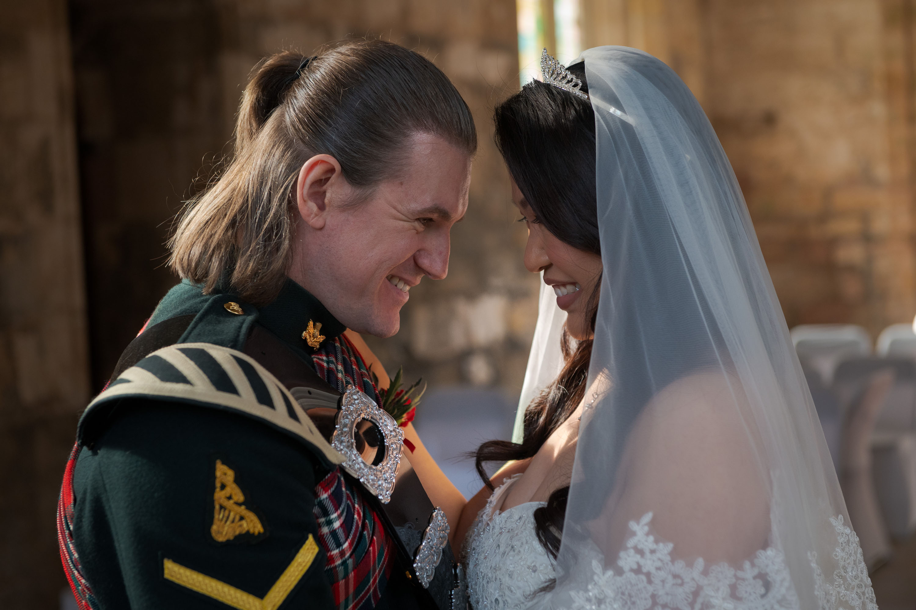 A bride and groom stand close together inside a historic stone building. The groom wears a dark green ceremonial uniform and a tartan sash. The bride wears a white lace wedding dress with a long sheer veil and a jeweled headpiece. Rows of chairs and stone walls are visible in the softly lit background.