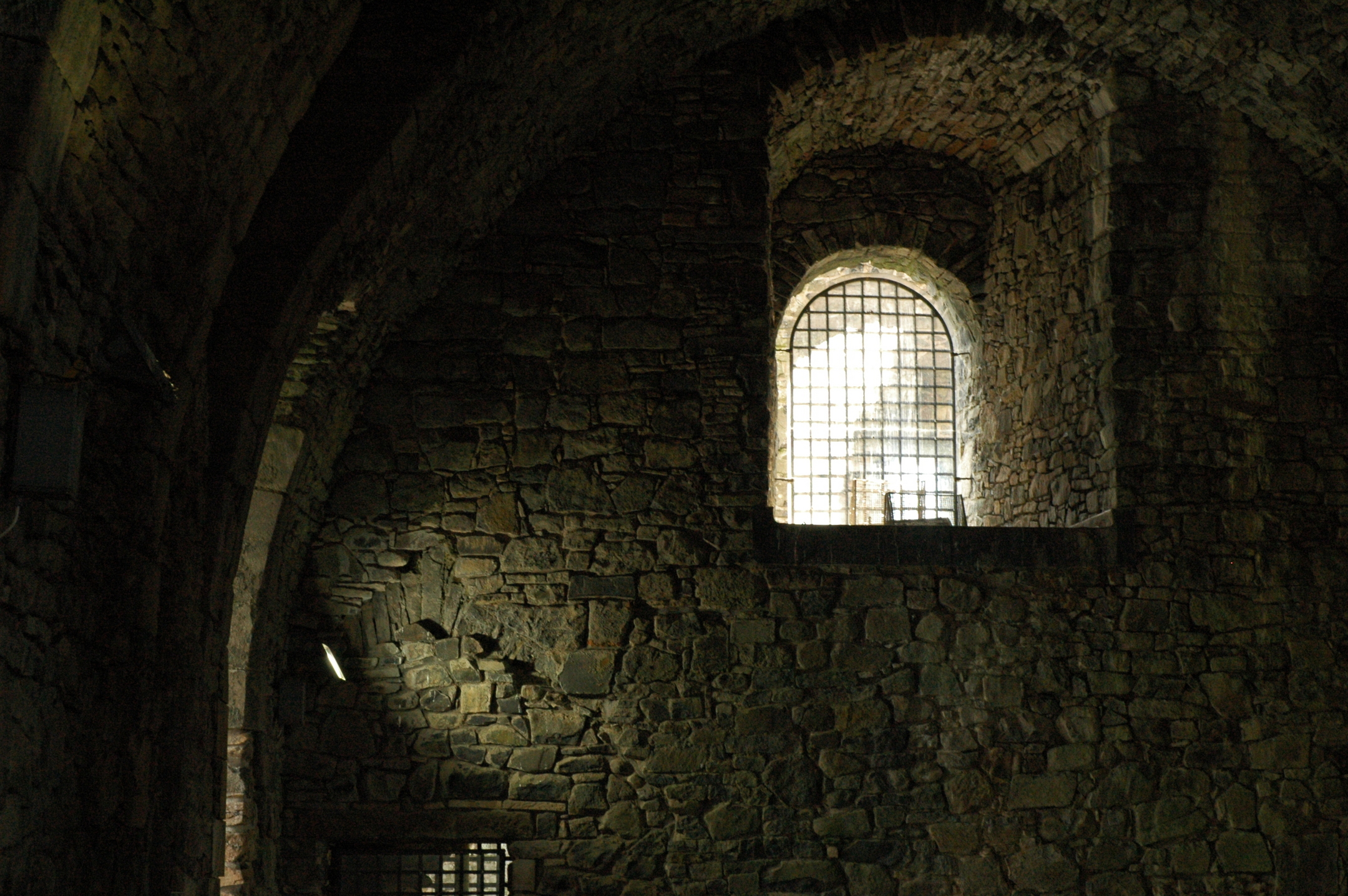 A dimly lit castle interior wall with a high arched window barred with iron grilles, catching bright light from outside.