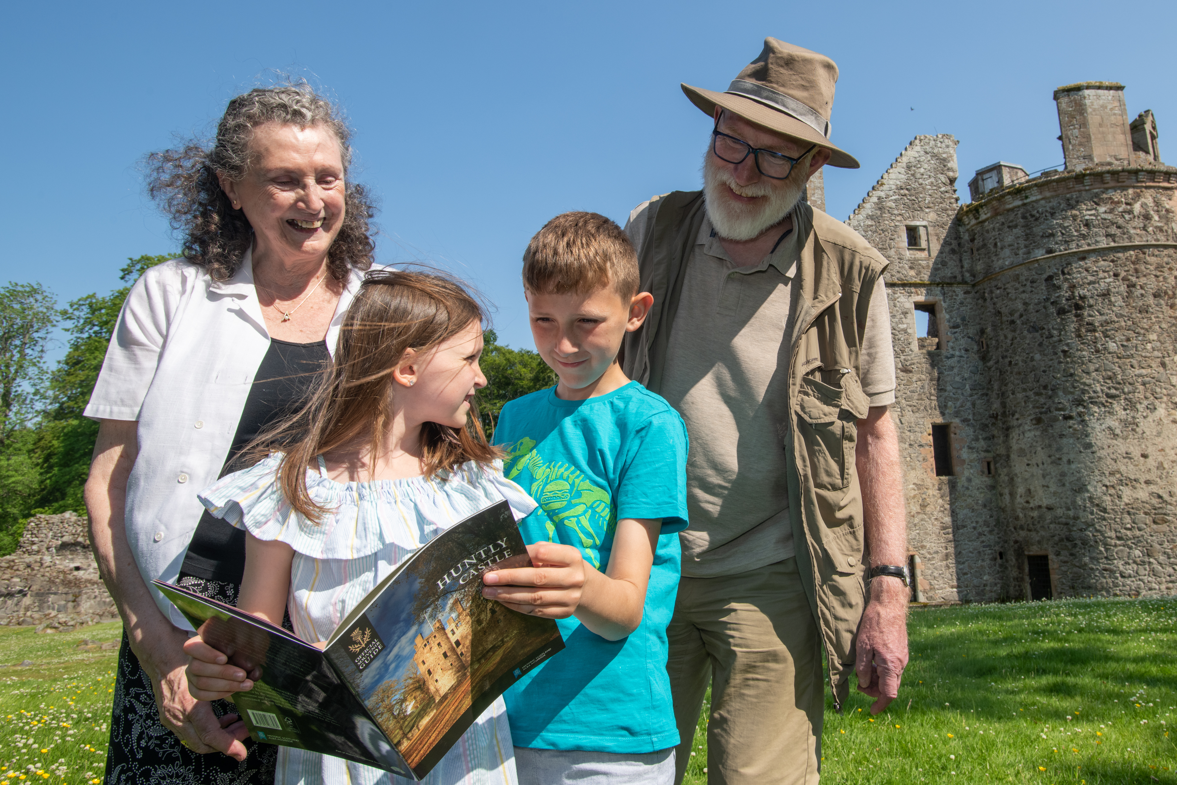 Two children smiling with their grandparents holding a guidebook. Caste run with grass under a blue sky is behind them.
