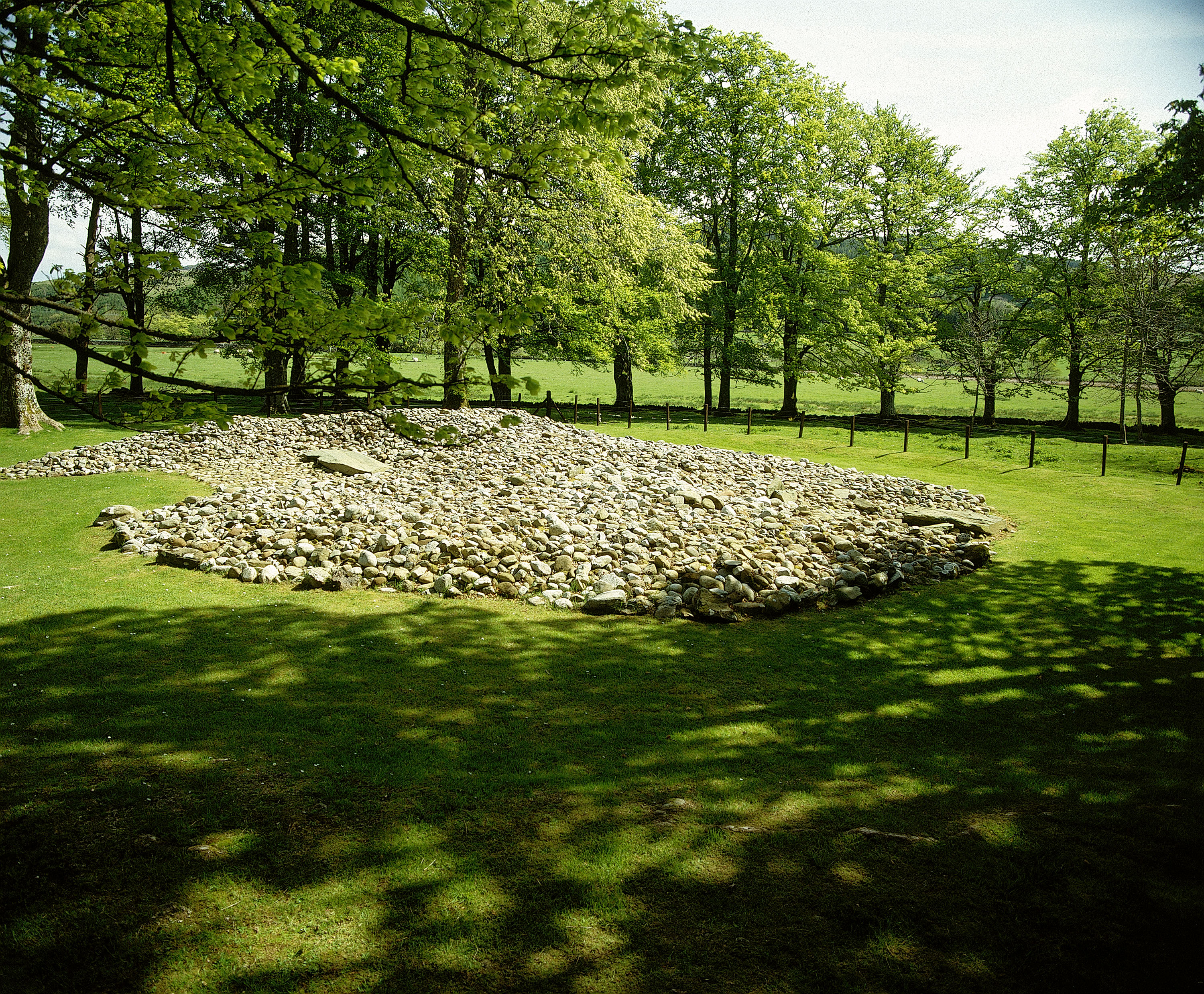 View of a low, circular cairn made of loosely piled stones set within a grassy clearing. The cairn has an uneven surface with a slightly raised central area. Tall trees with dense green foliage form a ring around the site, and sunlight filters through the branches, casting dappled shadows across the grass in the foreground. Open fields can be seen through the gaps in the trees.