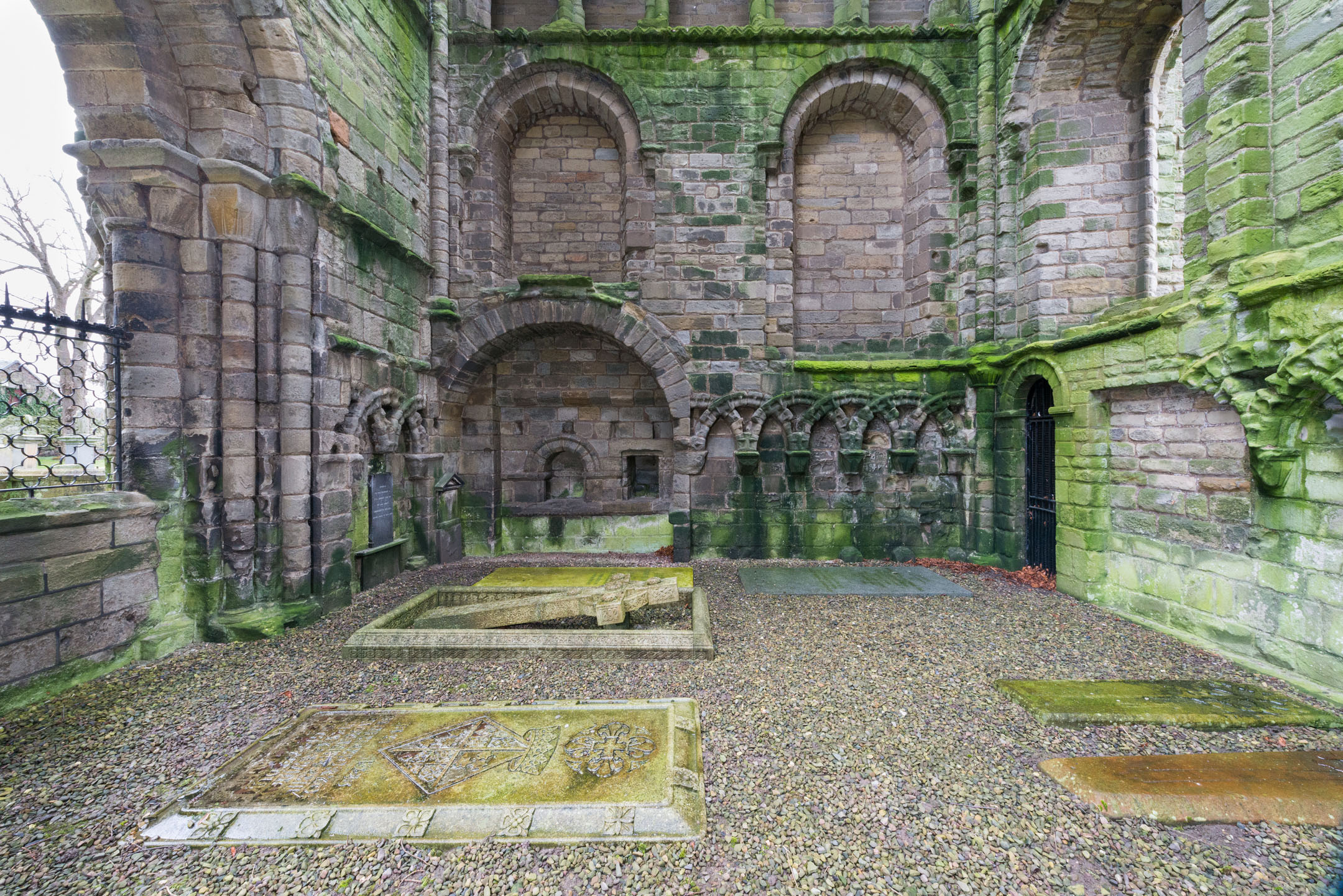 Interior wall with arcaded arches and multiple tombstones on gravel floor.