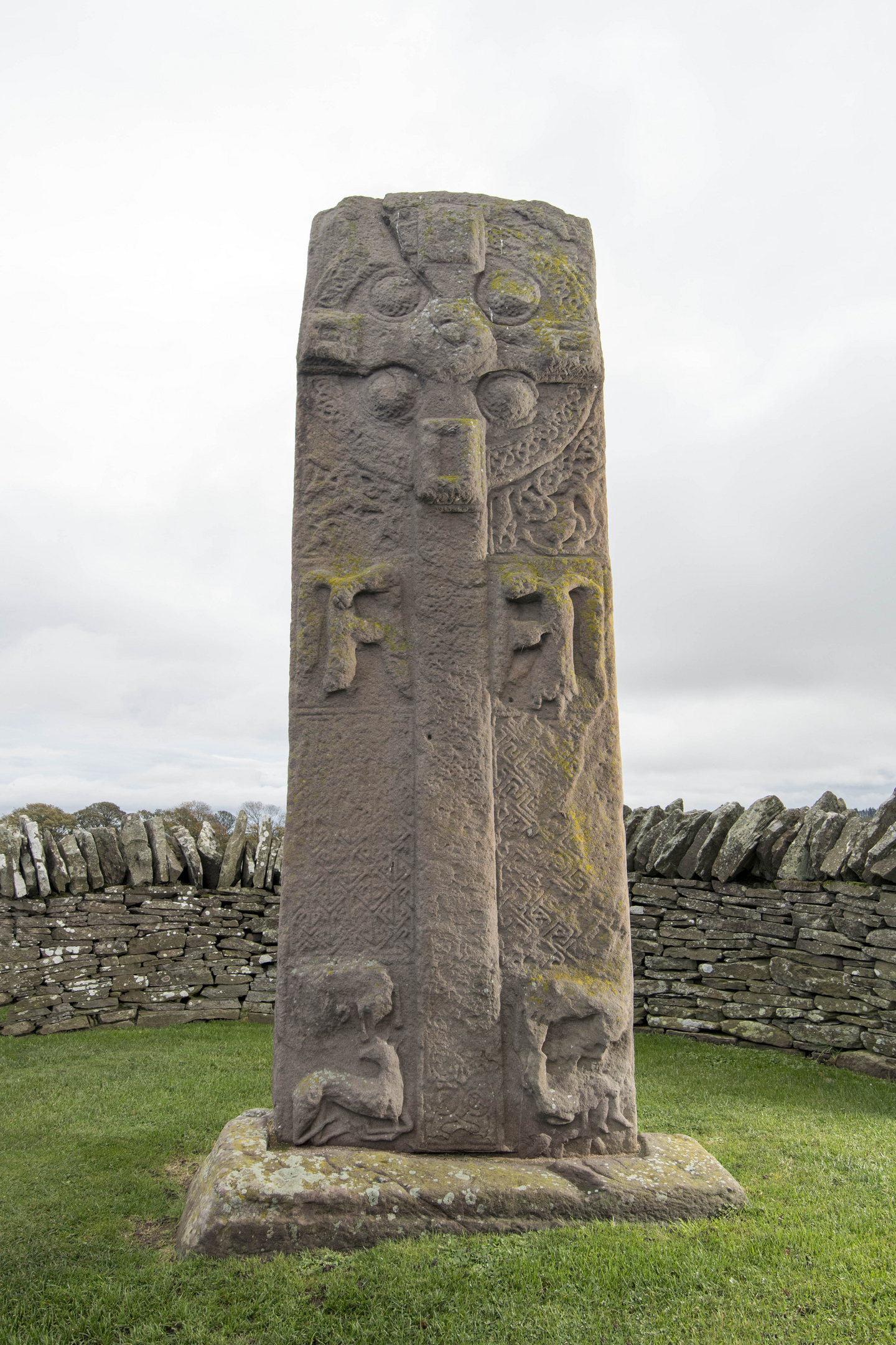 Sculptured stone with cross shape standing in grassy area with low wall behind