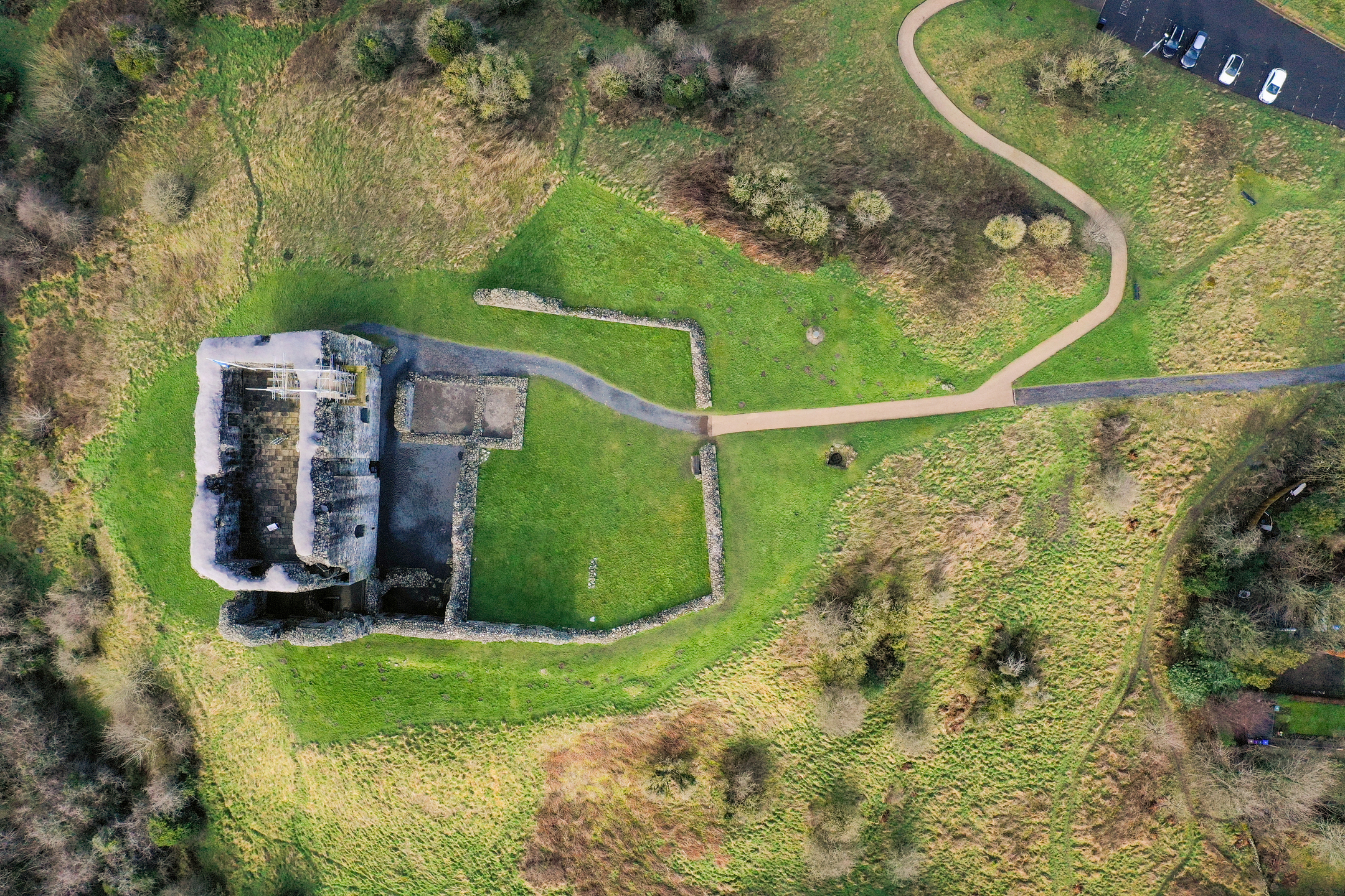 Top-down aerial view of the castle ruins and surrounding grassy area, with footpaths and a car park visible nearby.