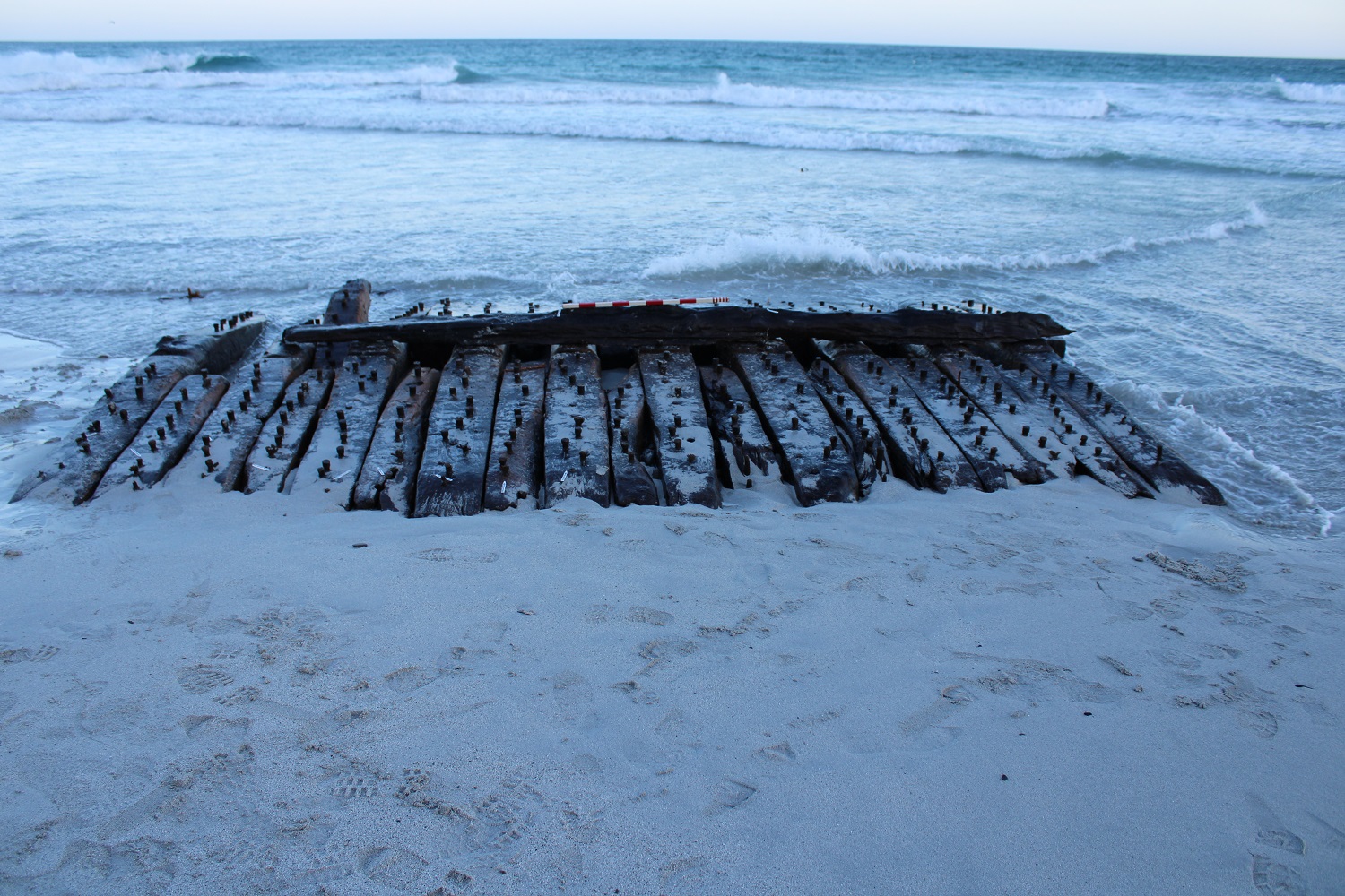 Old wooden timbers with rusty bolts partially buried in sand at the edge of a calm ocean shore with gentle waves.