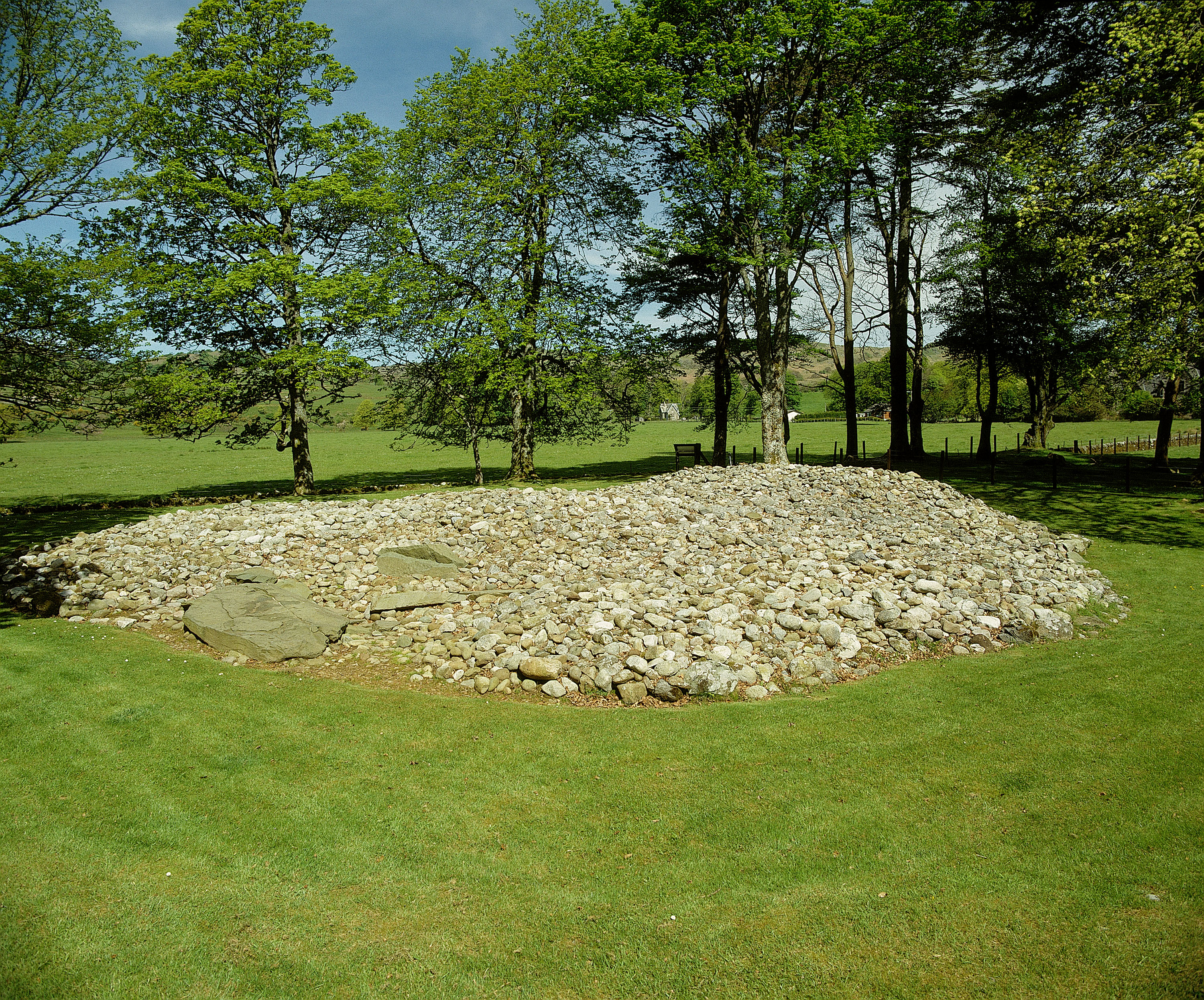 A low, circular cairn made of hundreds of loose stones sits in the middle of a grassy clearing surrounded by tall trees. The stones form an uneven mound with a slightly raised central area. Sunlight filters through the leafy branches overhead, casting dappled shadows across the grass in the foreground. The tree line encloses the site on all sides, giving the scene a sheltered, woodland setting.