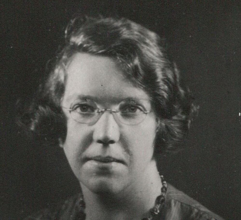 Black and white headshot portrait of a person wearing wire-rimmed glasses and a beaded necklace over a dark top.