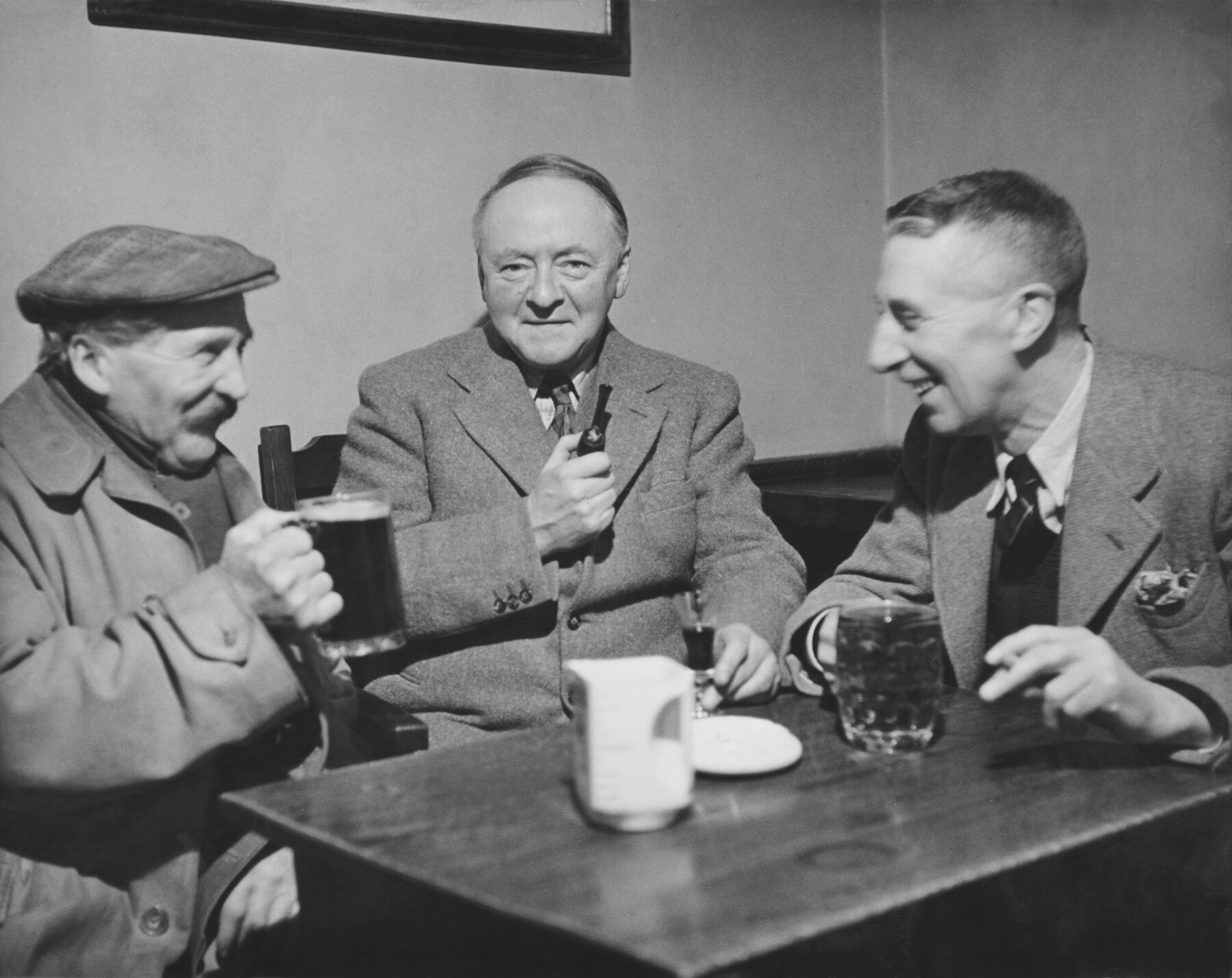 Black and white photo of three people seated at a table inside a public house. All are drinking and smiling, as if sharing a joke, and the person in the centre is looking at the camera and holding a pipe.