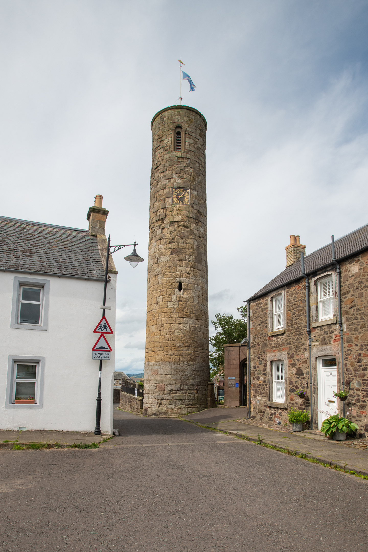 A tall view of Abernethy Round Tower rising at the meeting point of two quiet village streets, with traditional houses on either side.