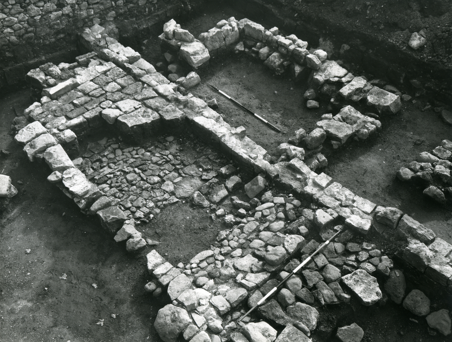 Black‑and‑white photo of an archaeological excavation showing low stone wall foundations arranged in rectangular sections. The ground inside the walls is uneven with scattered stones, and two measuring rods lie across the exposed structures.