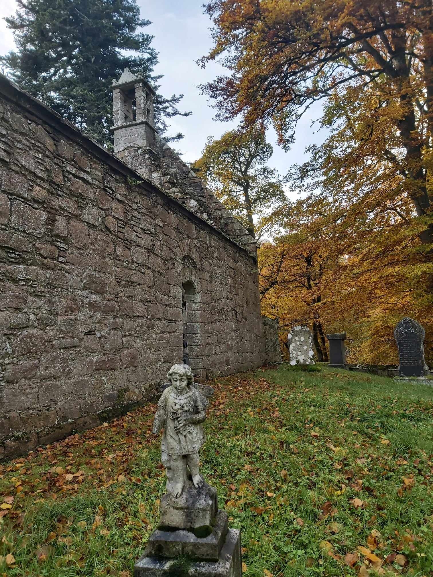 small stone statue next to the wall of a stone chapel, with autumn leaves scattered around the grass