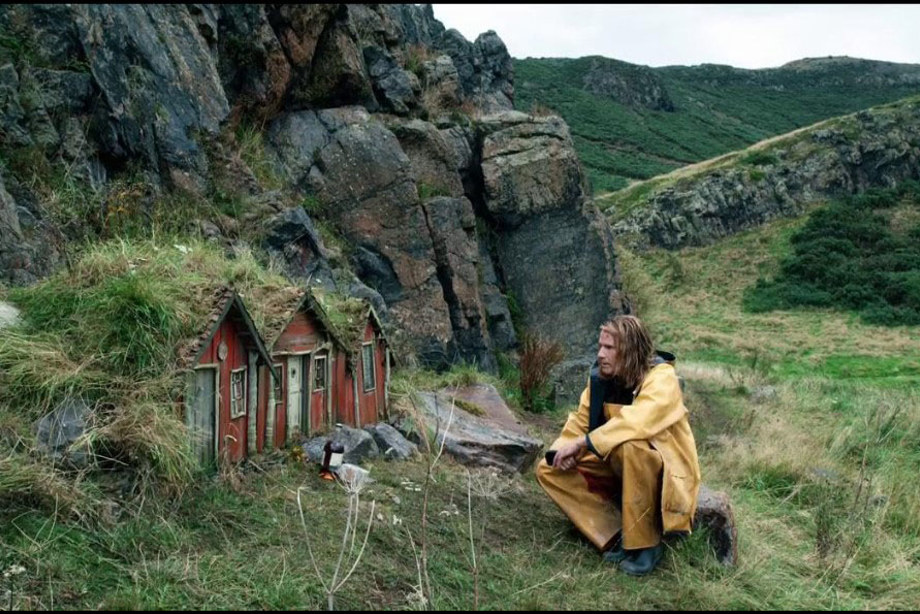 A man with long damp hair and bright yellow overalls and matching jacket, looking sombre sitting on a rock with a red miniature house opposite him 