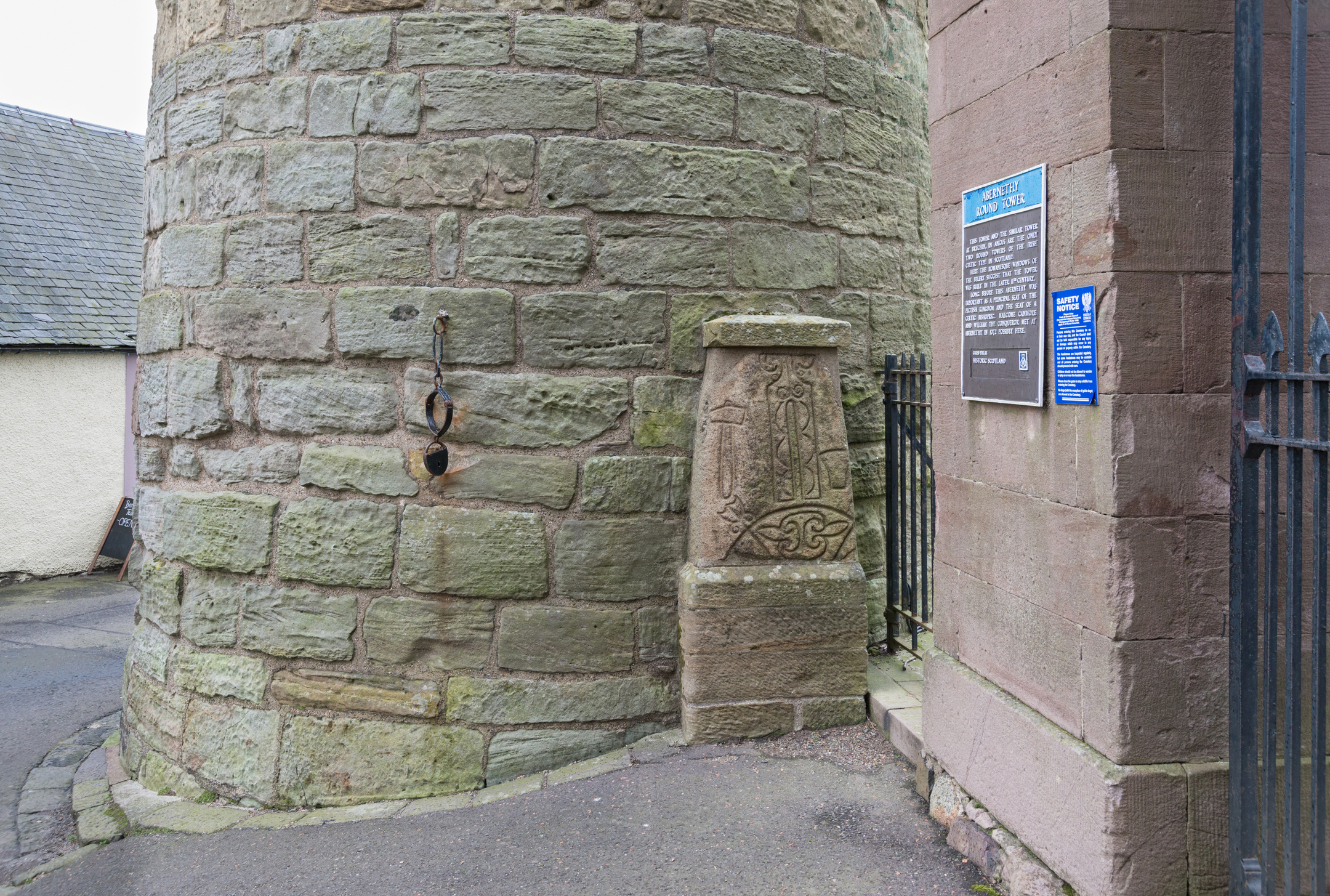 The lower section of Abernethy Round Tower showing its rough stone exterior, a carved Pictish symbol stone set against the wall, and a metal gate and information sign to the right.
