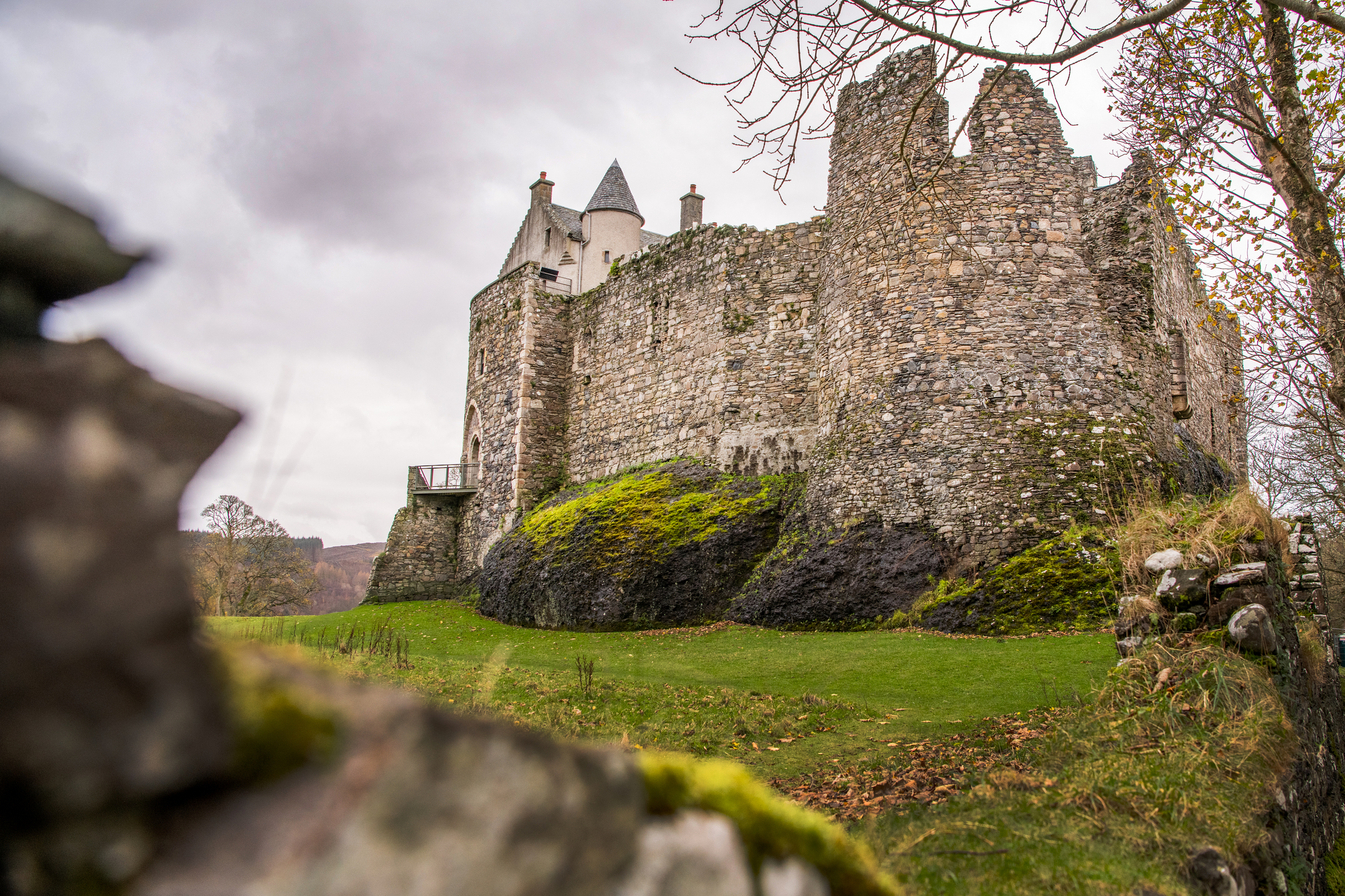 A moss‑covered stone castle wall rising from a rocky base, with autumnal trees and cloudy skies in the background.