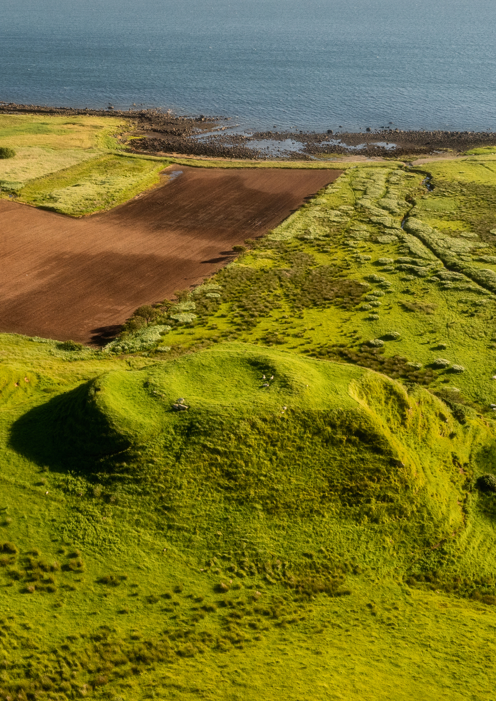A raised mound with ridge edges, there are fields and the sea in the background.
