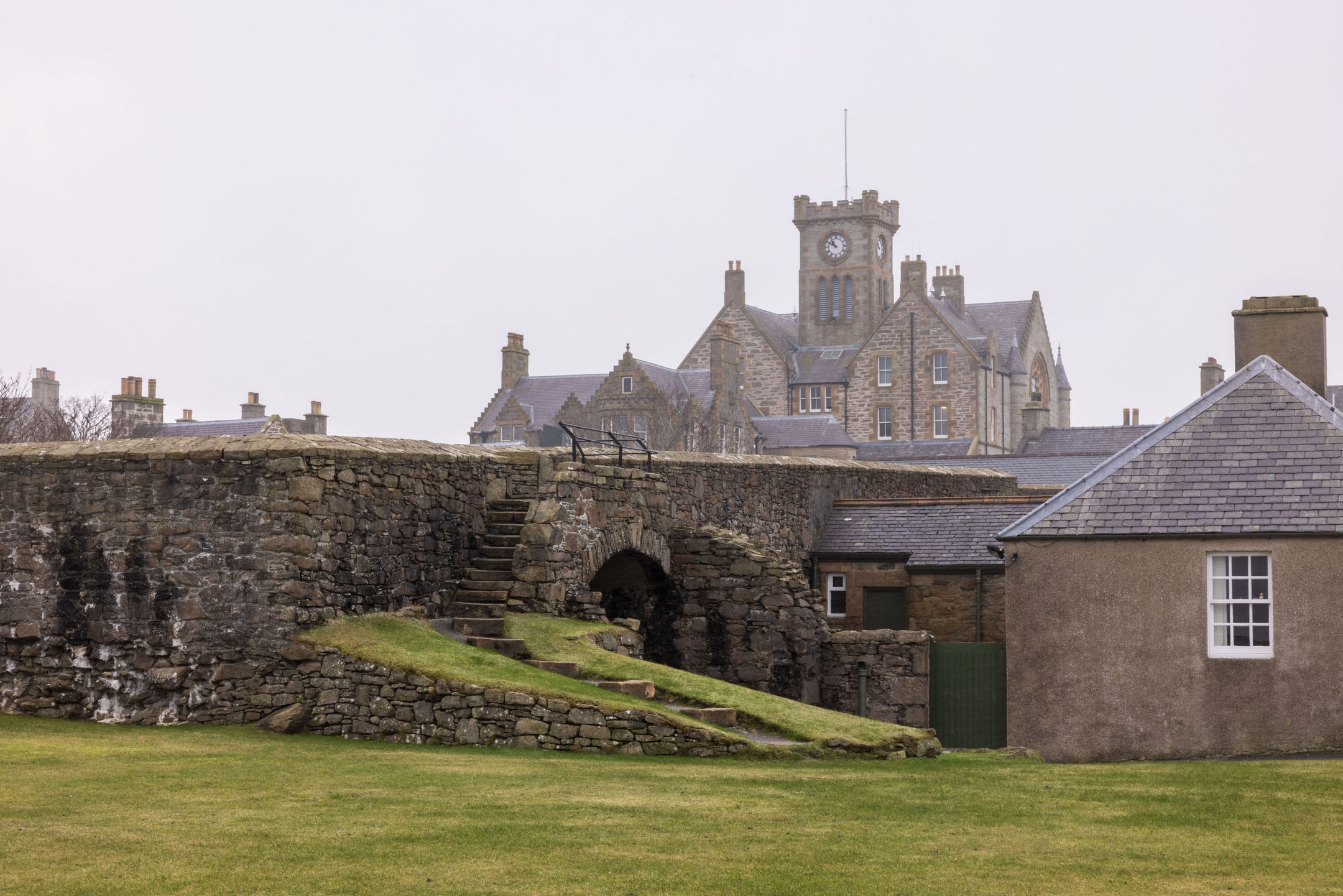 Grass bank and stone arch with steps leading up, town clock tower beyond.