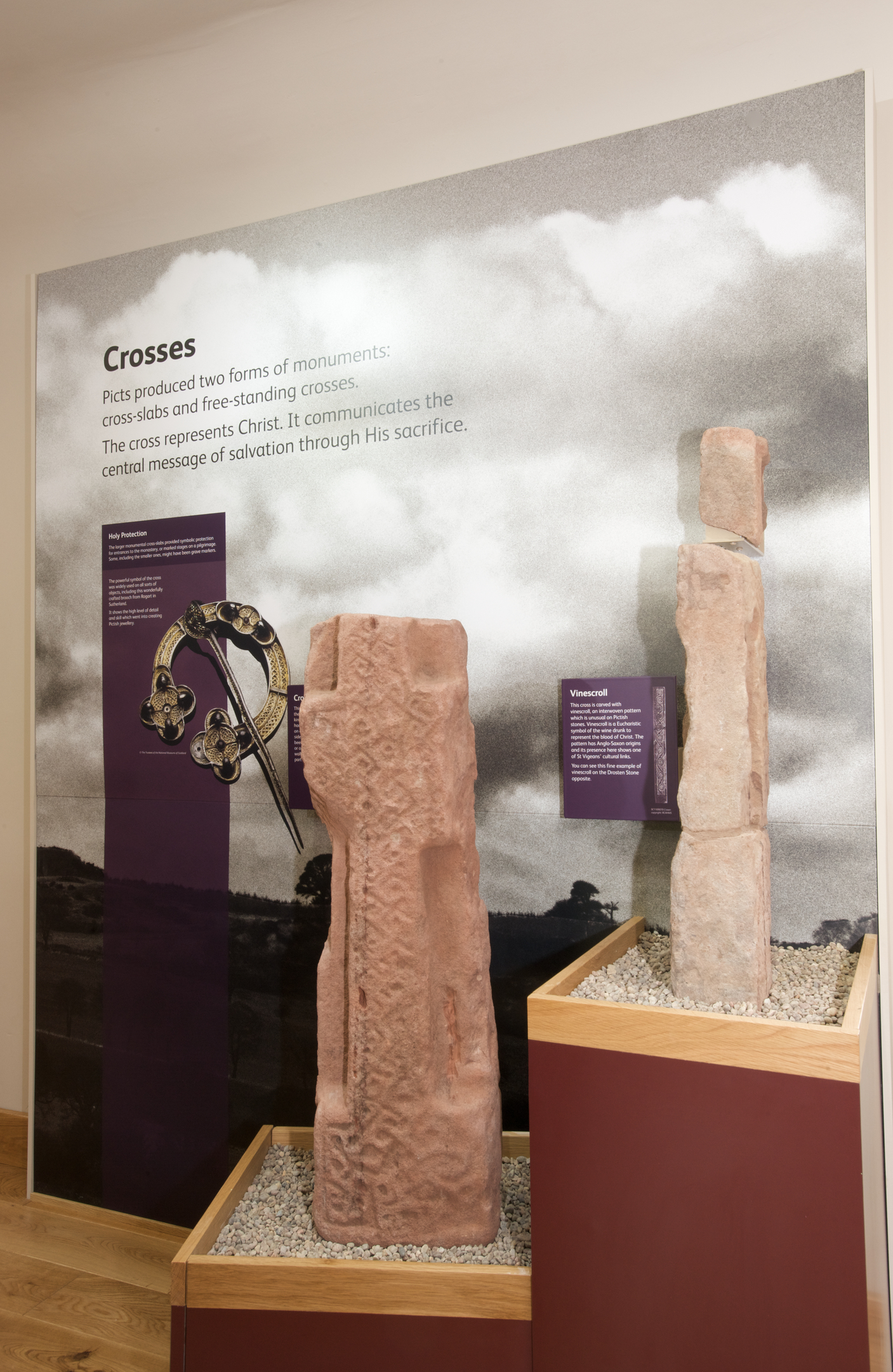 Panel titled “Crosses” with text about Pictish cross‑slabs, accompanied by two standing cross‑slabs carved with interlace patterns. Smaller interpretation boards are positioned beside the stones.