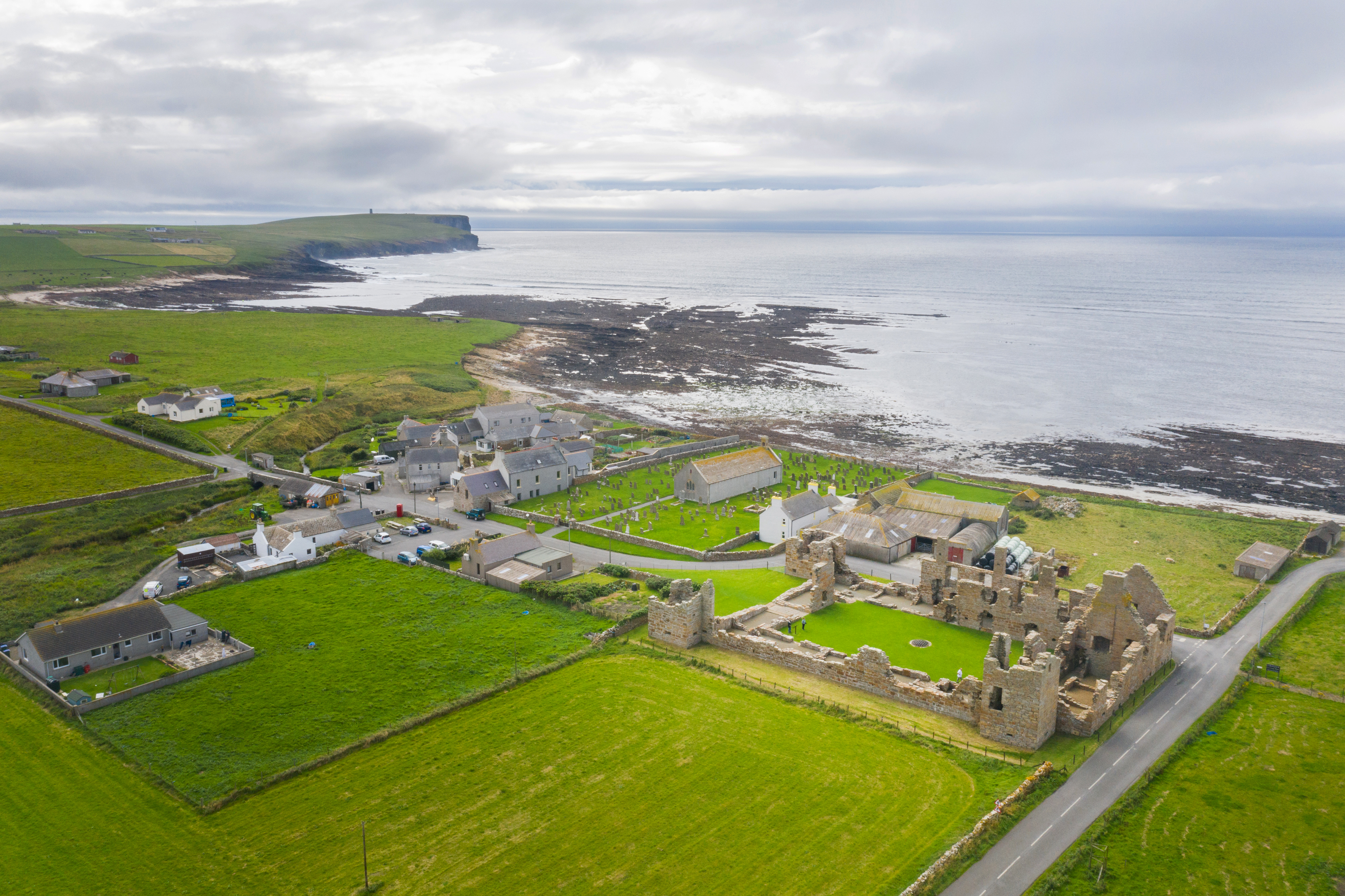 A drone shot showing Earl’s Palace in the centre of a small settlement near the Orkney coastline. Green fields, scattered houses, and the rocky shore stretch toward the sea beneath a cloudy sky.