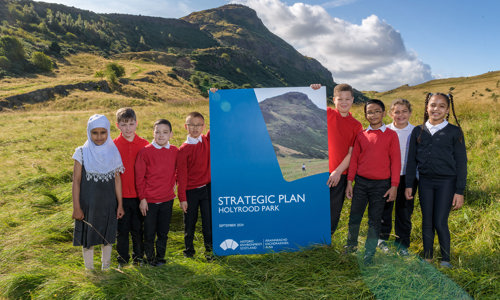 Eight children in red and black school uniforms stand on a grassy bank with a large grassy hill behind them. In the middle of them is a larger than life copy of the Strategic Plan for Holyrood Park. The sky is blue and the sun shining.