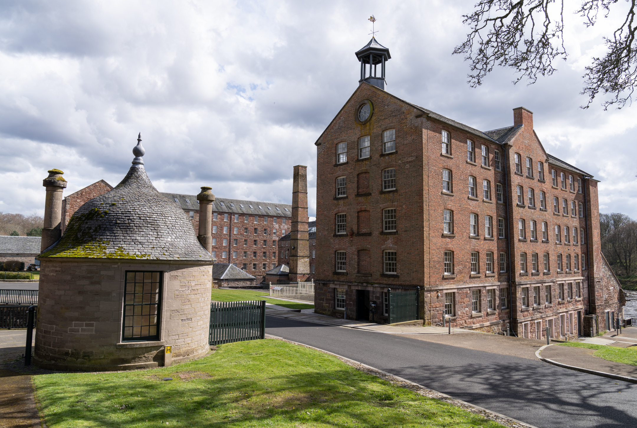 A cluster of multi‑storey brick mill buildings, a chimney, and smaller outbuildings form a historic industrial complex framed by lawns and pathways.